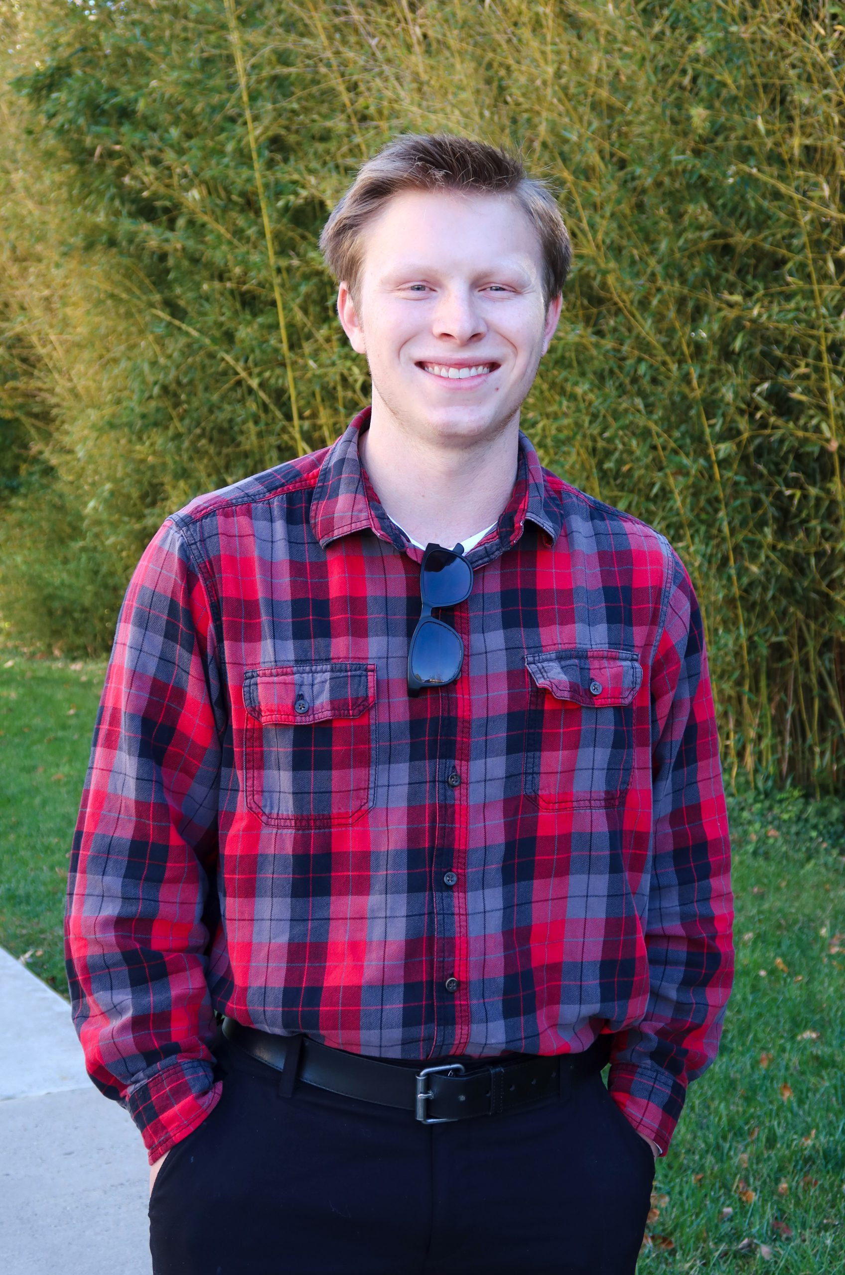 Outside on a sunny fall day, NOVA student Jacob Clasen smiles for the camera. He wears a red, blue and black plaid shirt, with dark sunglasses hanging at the collar.