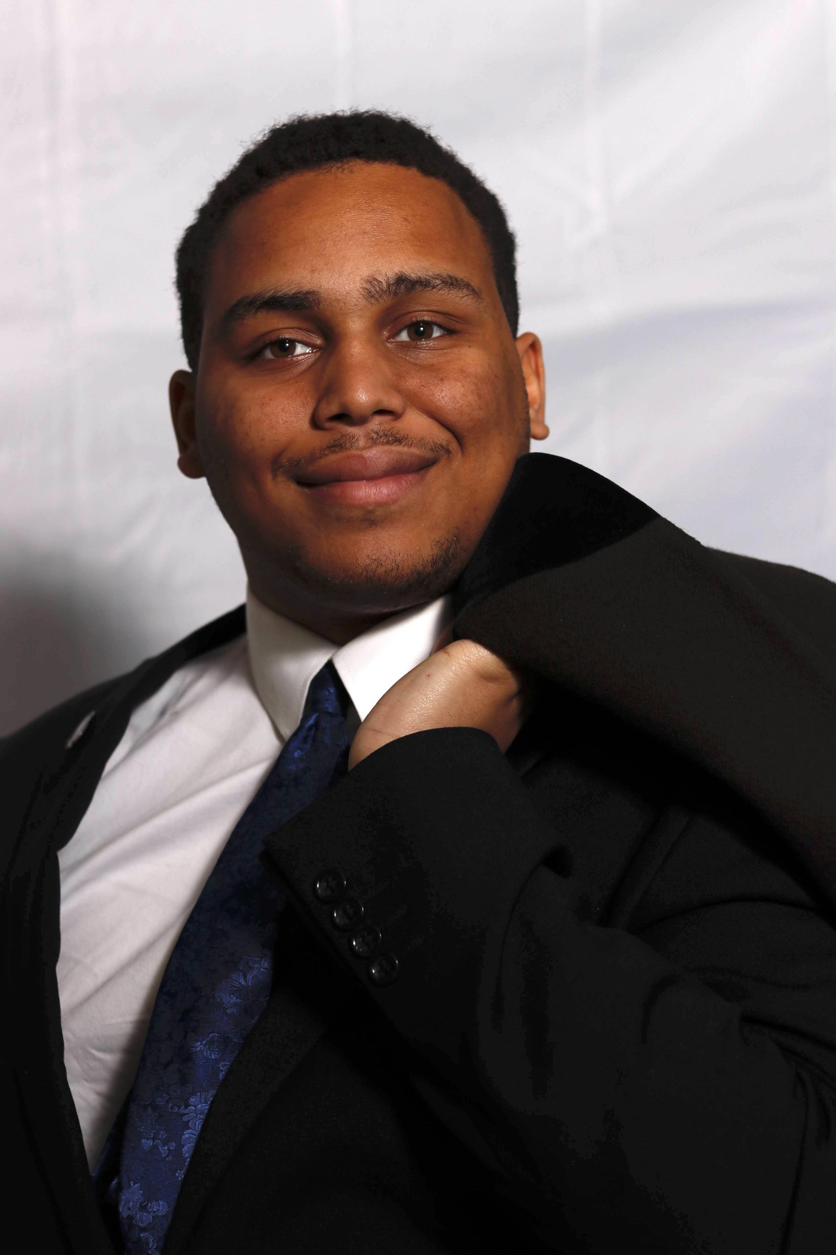 Josiah Greene, wearing a dark suit, white collared shirt, and dark tie, smiles for the camera.