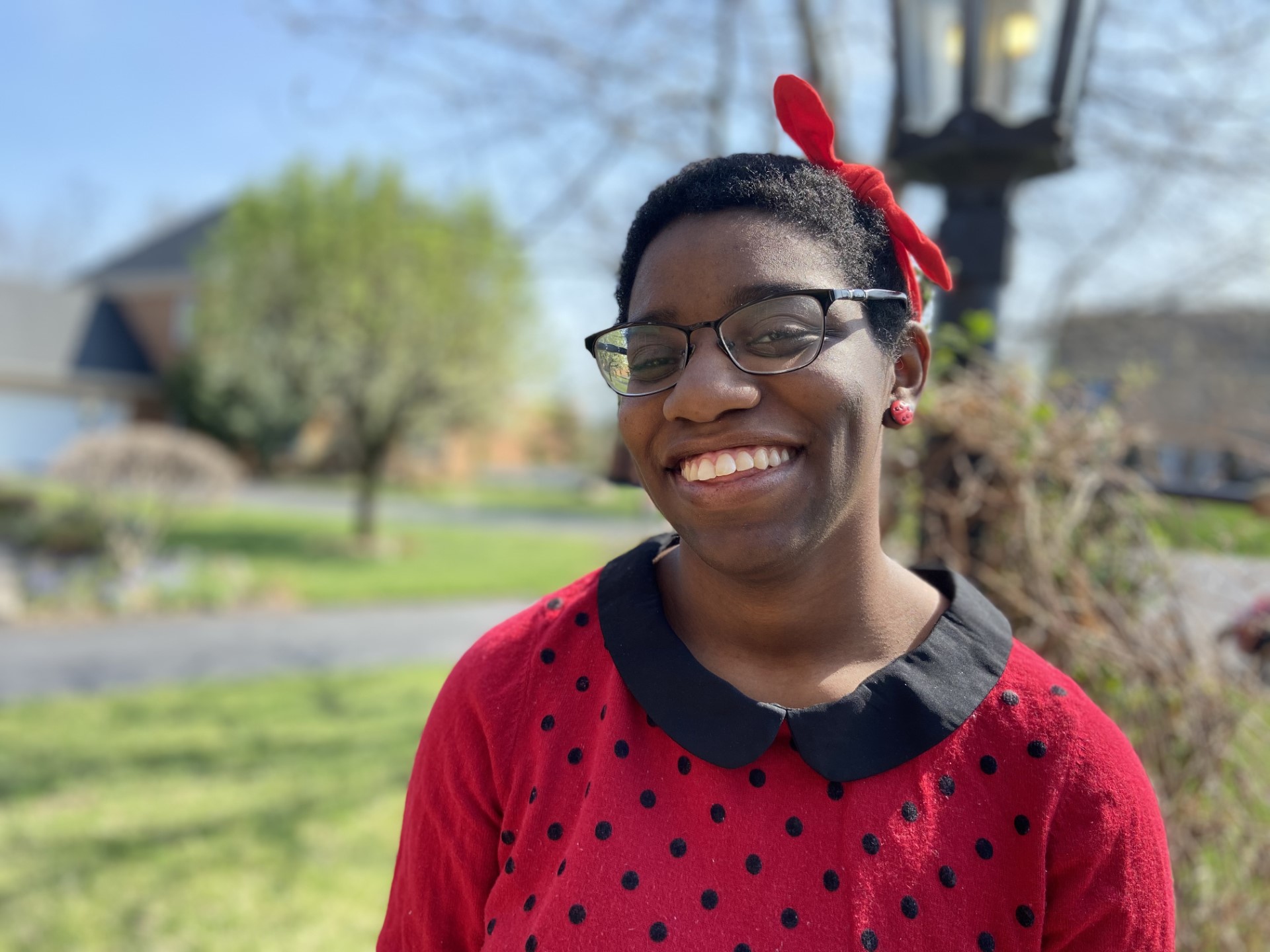 Kylee "Lee" Kennerly smiles for the camera on a beautiful sunny day. Lee wears a red top with black polka dots and black collar, dark eyeglasses, and a red bow in their hair.