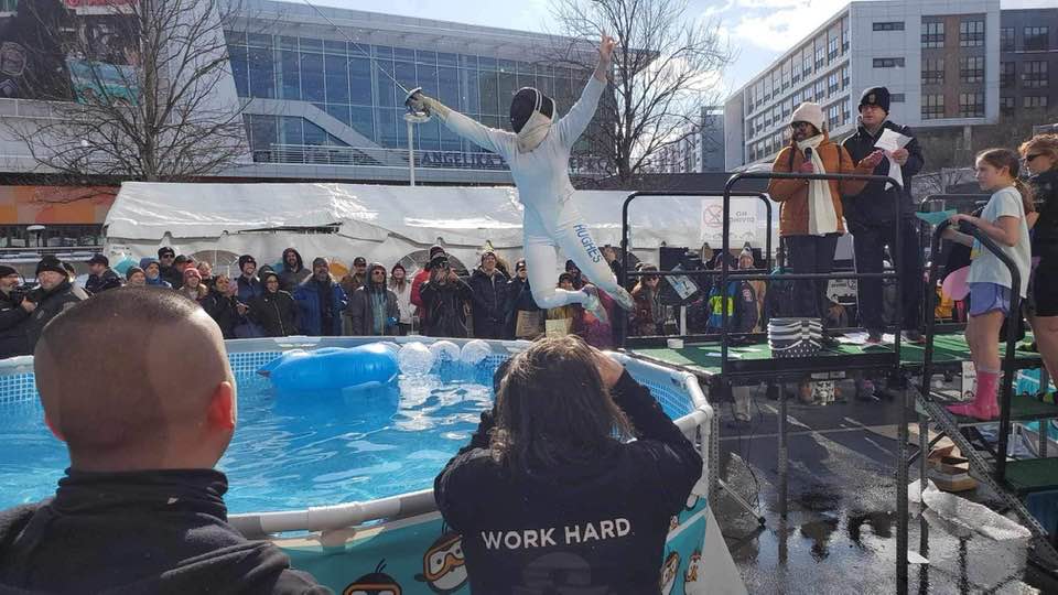 Erica Hughes, dressed in full fencing attire and holding her fencing foil, jumps victoriously into an ice-cold pool set up in the common space of the Mosaic District in Falls Church. A crowd of onlookers cheer her on. Hughes is helping to raise money for Special Olympics.