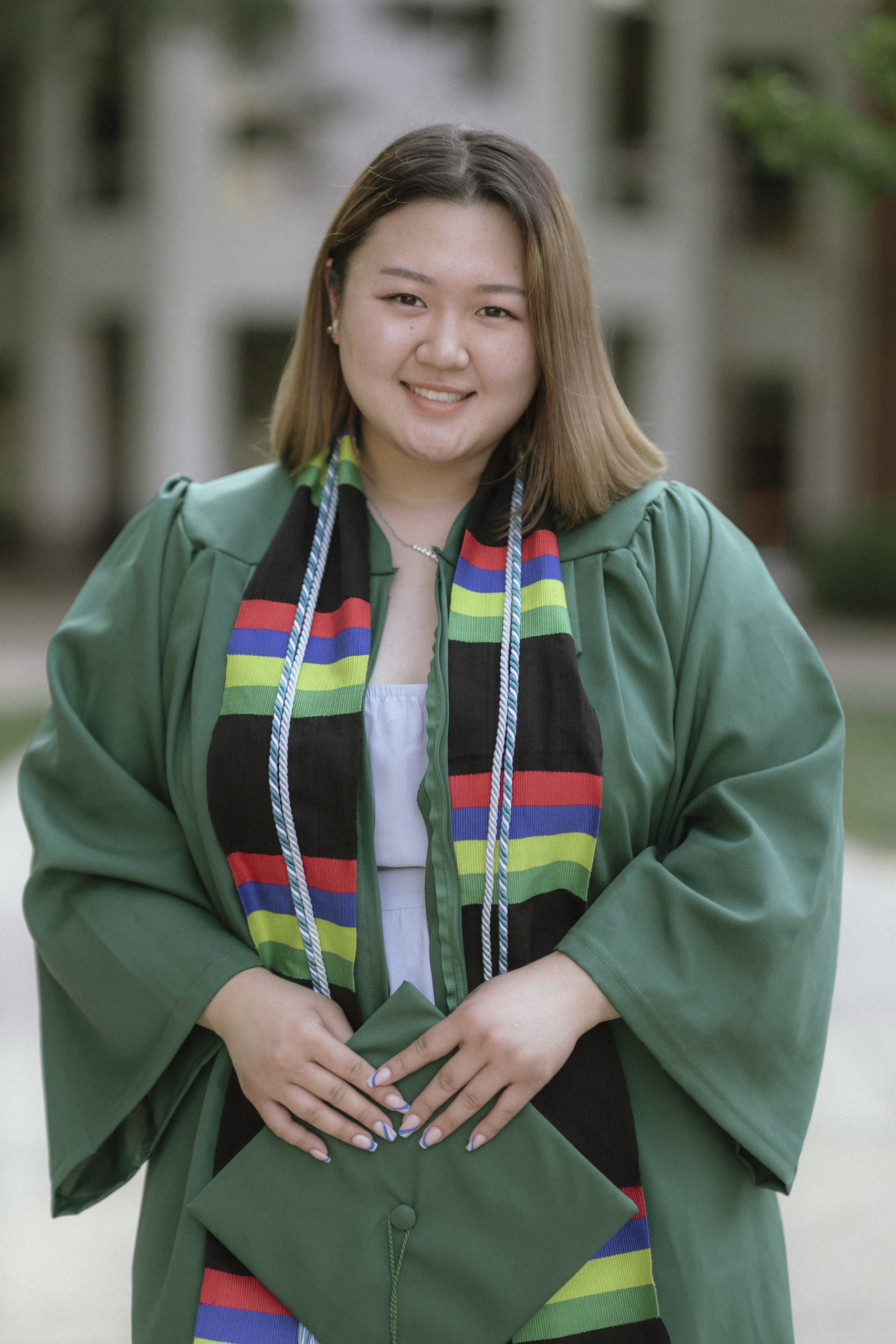 ADVANCE alumna Hannah Choi, in her green NOVA graduation gown, smiles for the camera while holding her green mortarboard graduation cap. She wears a colorful stole and two graduation cords.