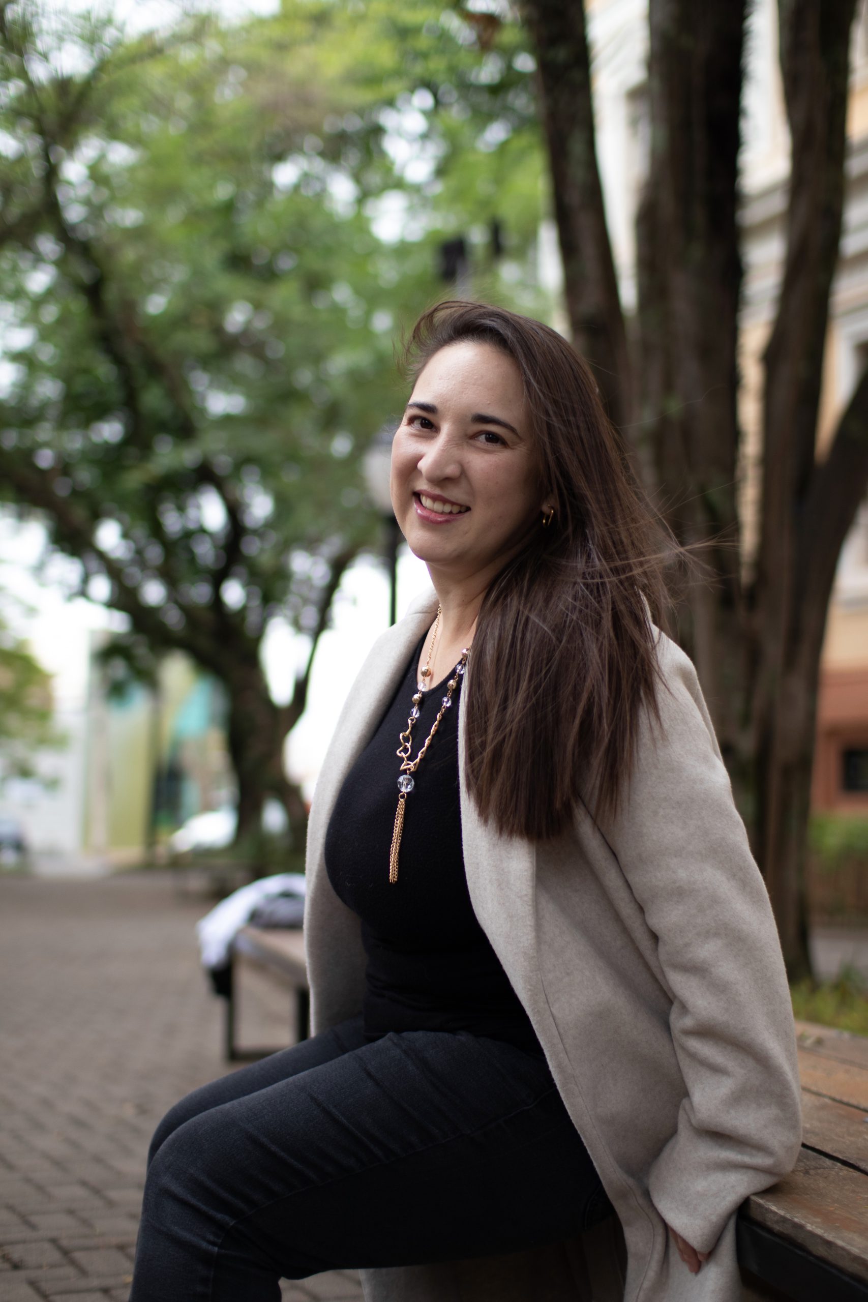 Bruna Mayen, with long dark hair swept to the side and a beige jacket, smiles as she sits on a peaceful bench outside a treed brick walkway.