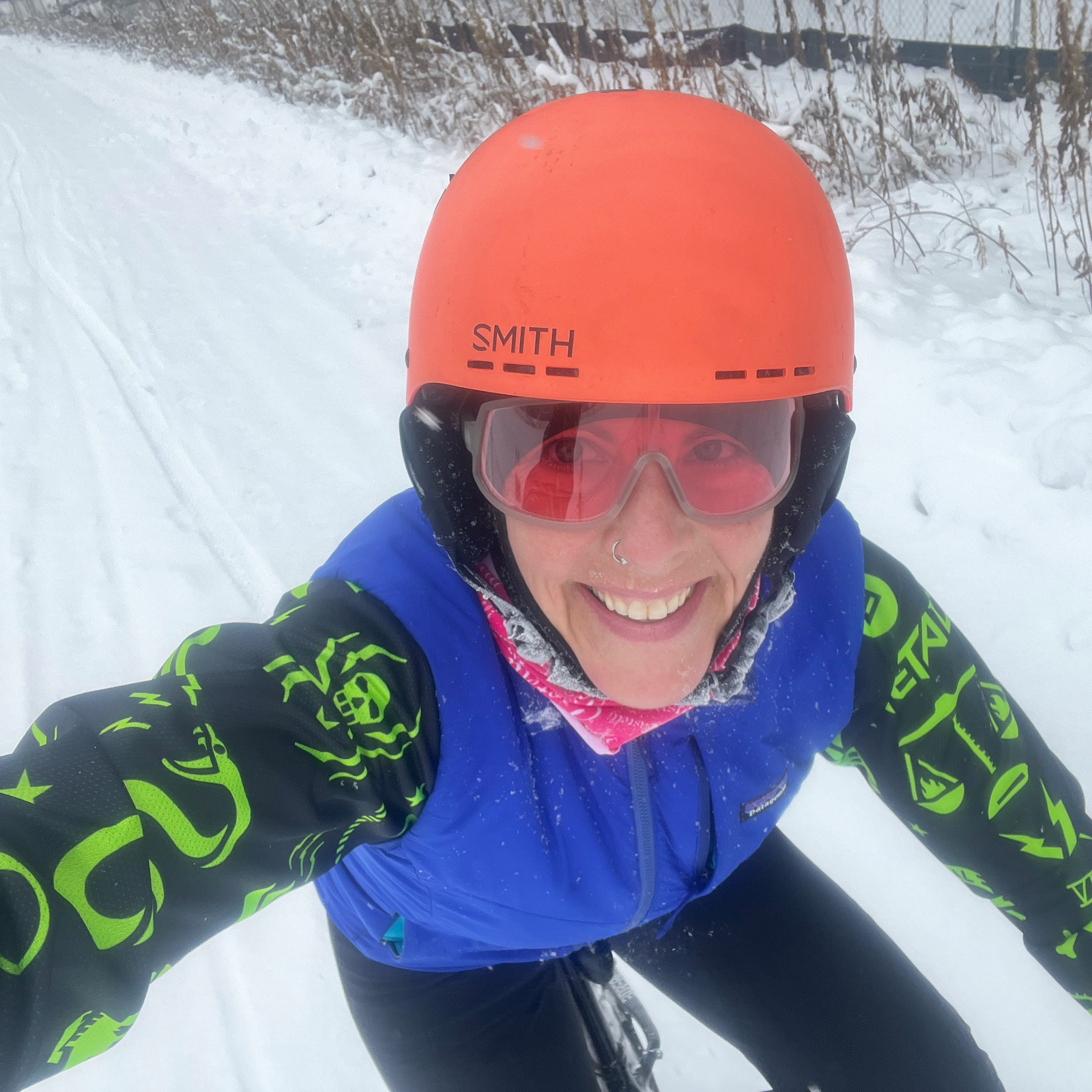 Artist Kate Fisher takes a selfie while snowboarding. She smiles for the camera while wearing an orange helmet and goggles, a blue snow vest, and a green and black printed shirt.