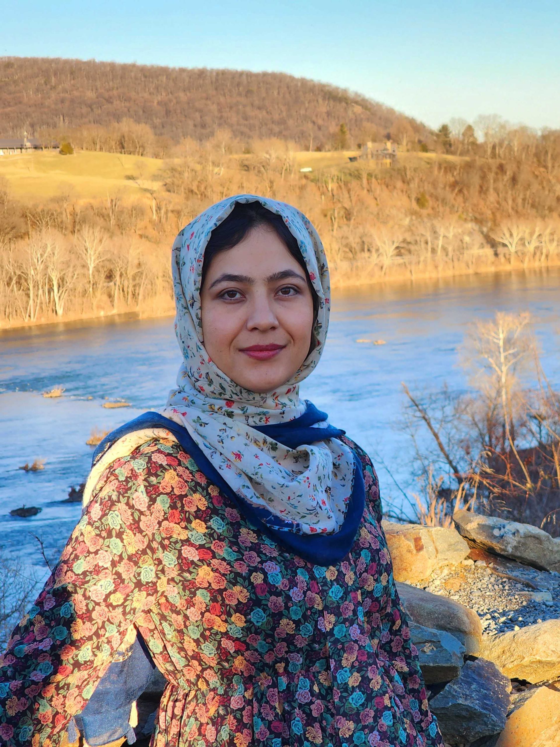 NOVA Nighthawk Manizha Khorram smiles for the camera against a natural backdrop of a flowing blue river on a bright winter's day. With bare trees on the riverbank in the background, she wears a floral long-sleeved dress and a printed headscarf.