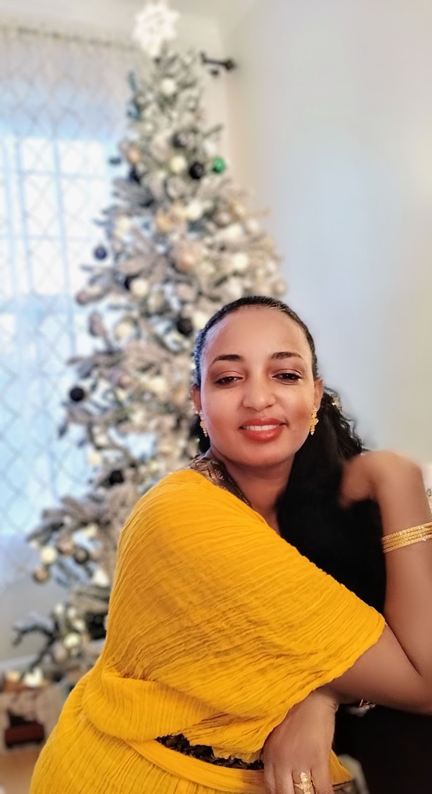 NOVA student Milka Tedla poses while smiling in front of her family Christmas tree. Milka, with long dark hair pulled back from her face, wears a crepey yellow dress and gold jewelry. Her living room is white, and her beautiful Christmas tree is also decorated in shades of winter white.