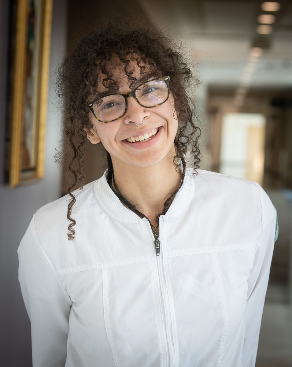 NOVA nursing student Maya Smith, wearing tortoiseshell glasses and a white nursing uniform, smiles for the camera at the Medical Education Campus.