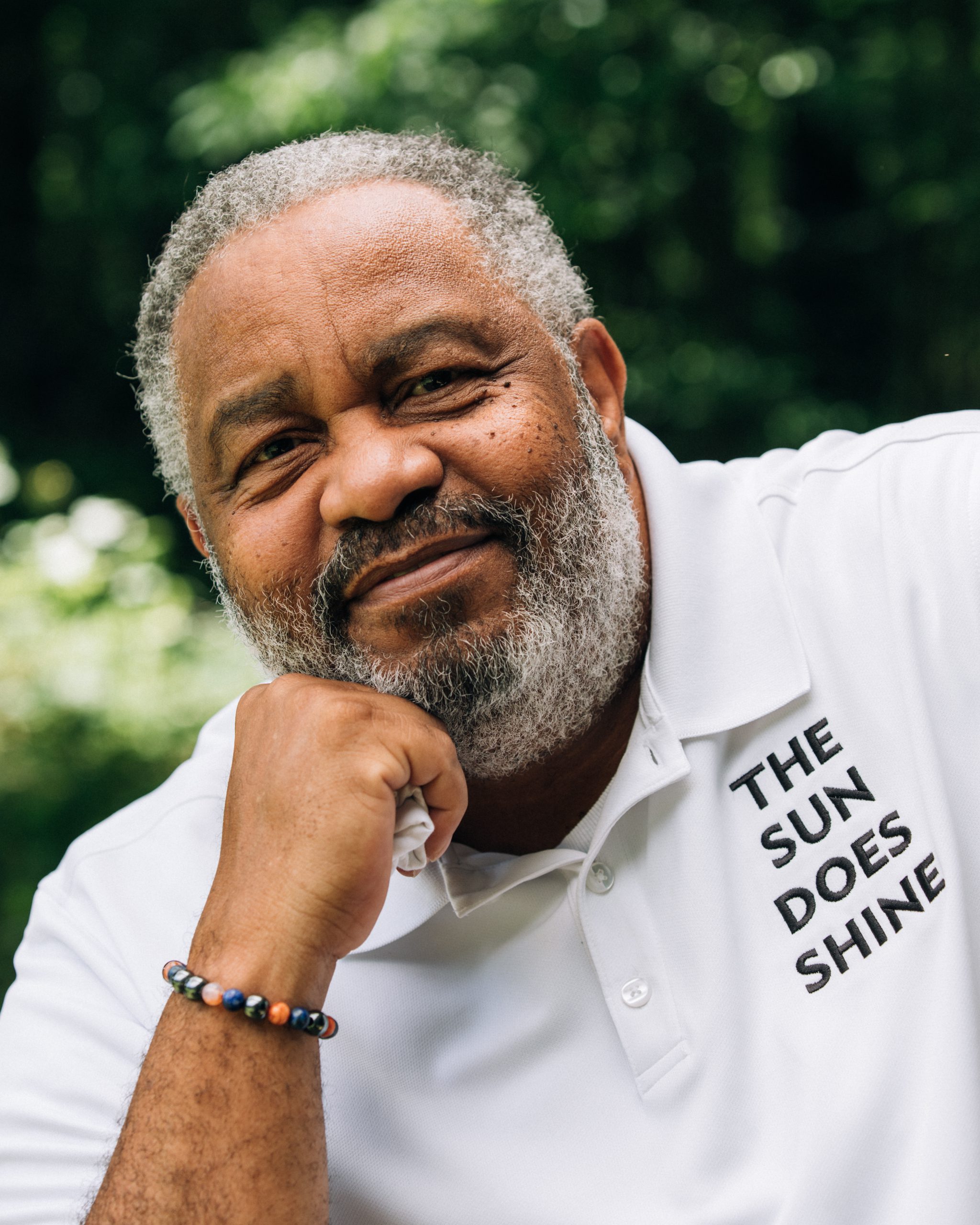 Anthony Ray Hinton smiles for a portrait outdoors with trees in the background. He wears a white polo shirt emblazoned with "The Sun Does Shine." Photo credit: Rob Liggins.
