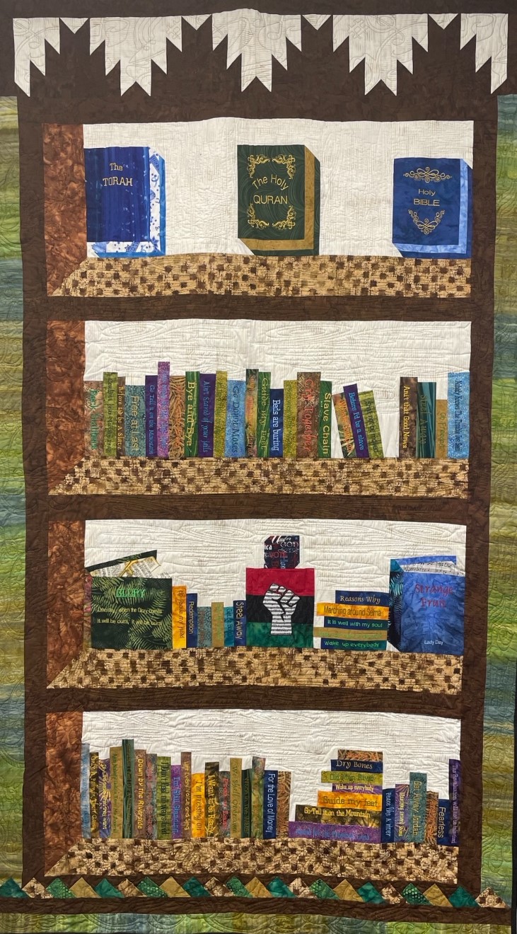 Freedom Bookshelf shows amazing workmanship with a grass green and blue wide striped background behind a quilted bookshelf with books on four shelves, a copy of the Torah, the Quran and the Holy Bible on the top shelf; books of black life and culture on the second and fourth shelves; boxes of sheet music, books and a pan-African flag on the third shelf.