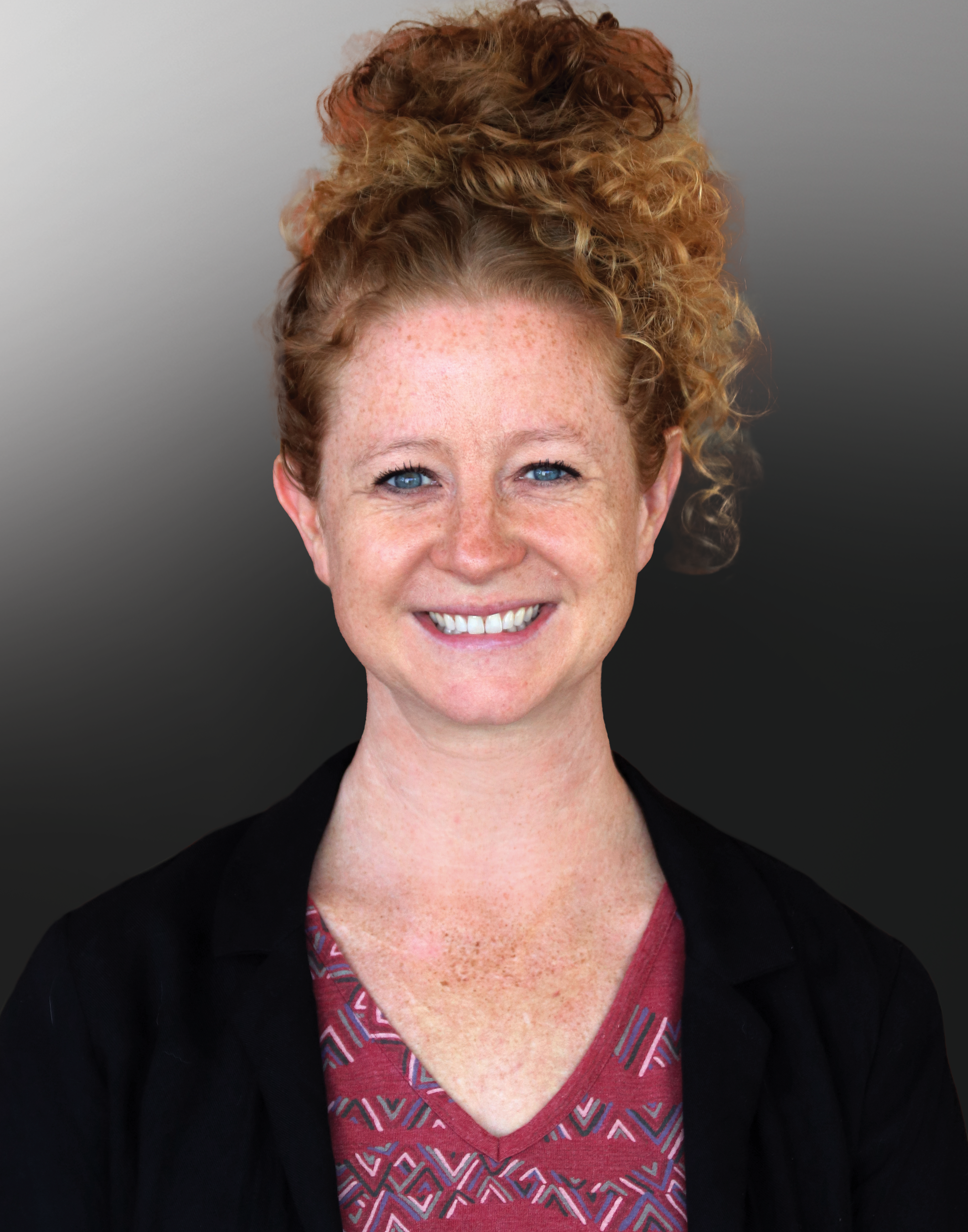 NOVA Professor Indigo Eriksen, with her hair arranged on the top of her head, smiles for the camera in her professional headshot. She wears a red patterned blouse under a black blazer. 