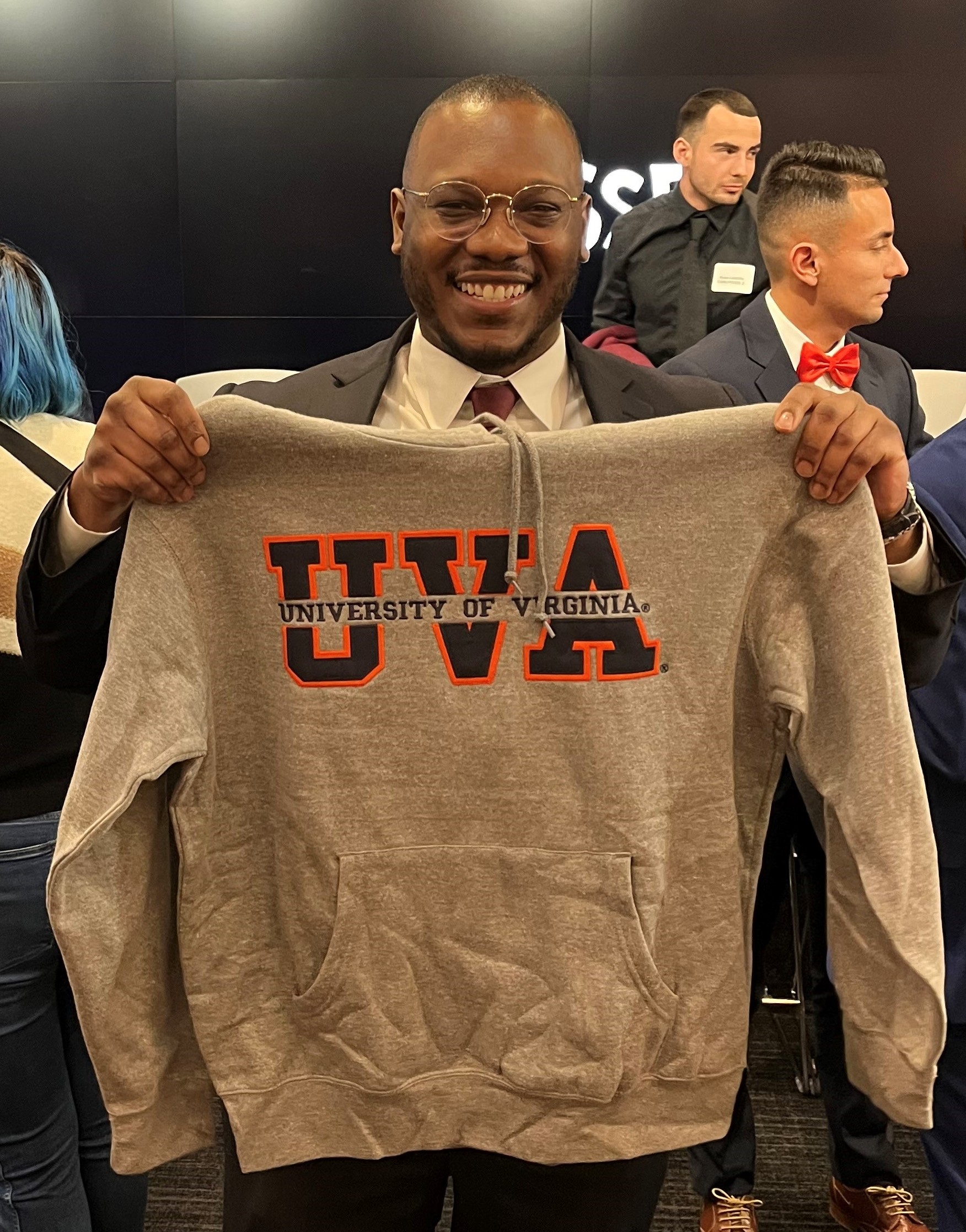 NOVA Nighthawk and Posse Veterans Program Scholarship recipient Justin Howard, wearing a tie and glasses, smiles for the camera while holding up a gray University of Virginia sweatshirt.