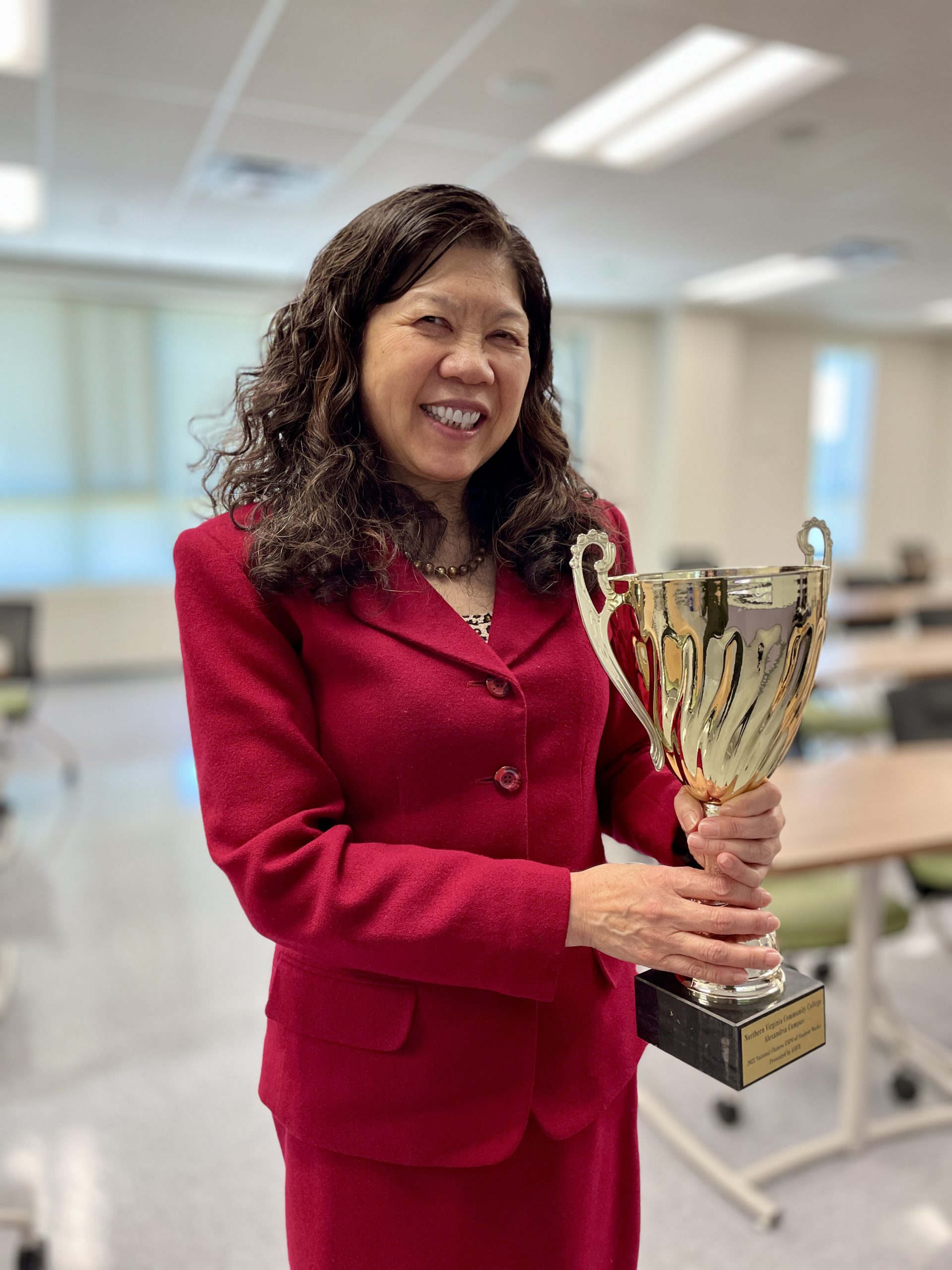 Professor Dali Tan, wearing a red skirt suit in an empty NOVA classroom, smiles for the camera while holding a large gold cup — one of her many teaching awards! 