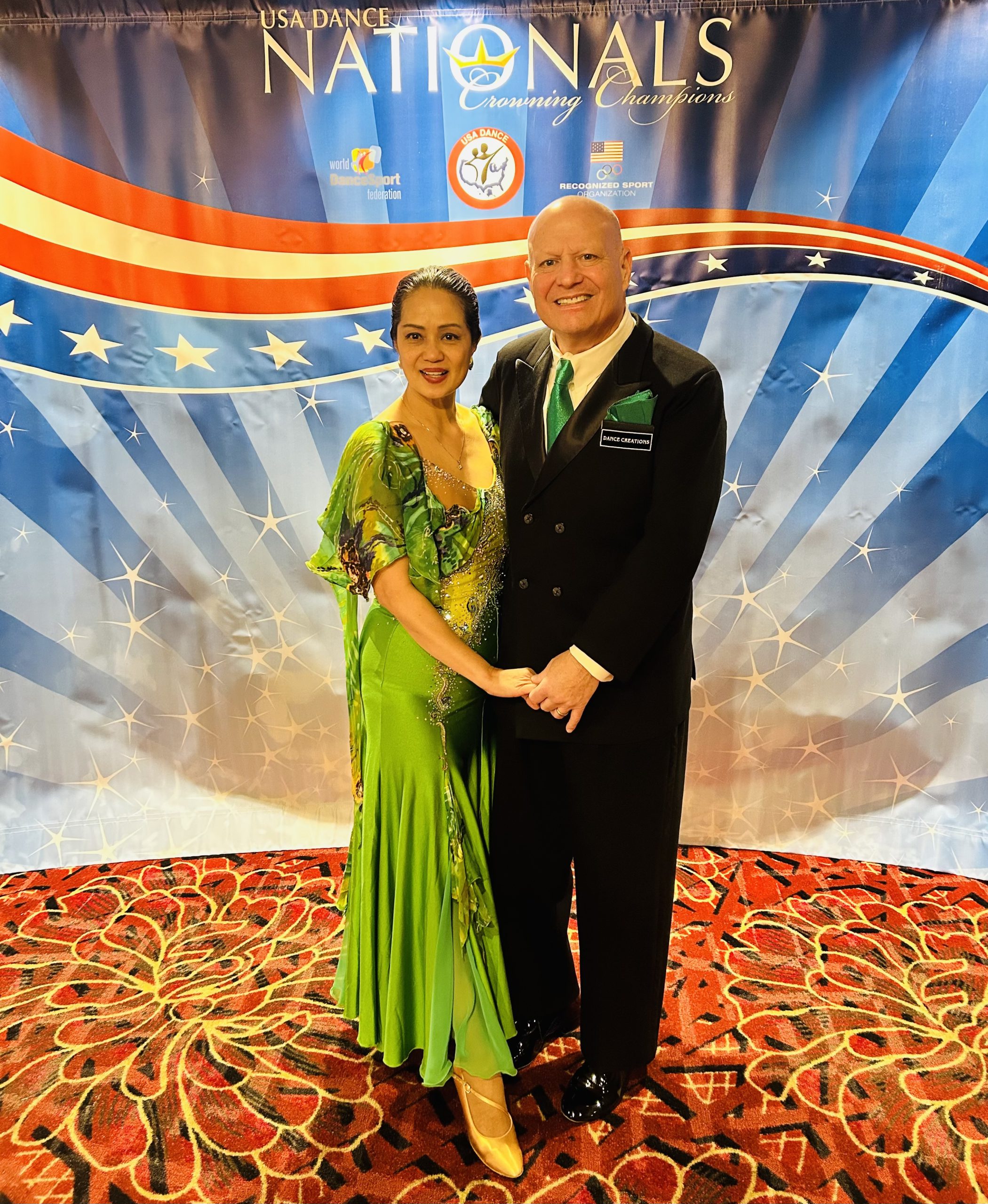 Photo of Alan Golombek wearing a black tuxedo and green tie and his wife (also dance partner), Thuy, wearing a flowing green gown. The couple stand in front of a red, white, and blue backdrop emblazoned with the words "USA Dance Nationals, Crowing Champions."