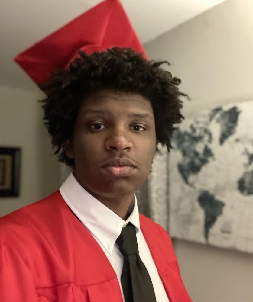 Muhanad Osman poses with his red cap and gown from high school graduation. In the background is a decorative black and white world map