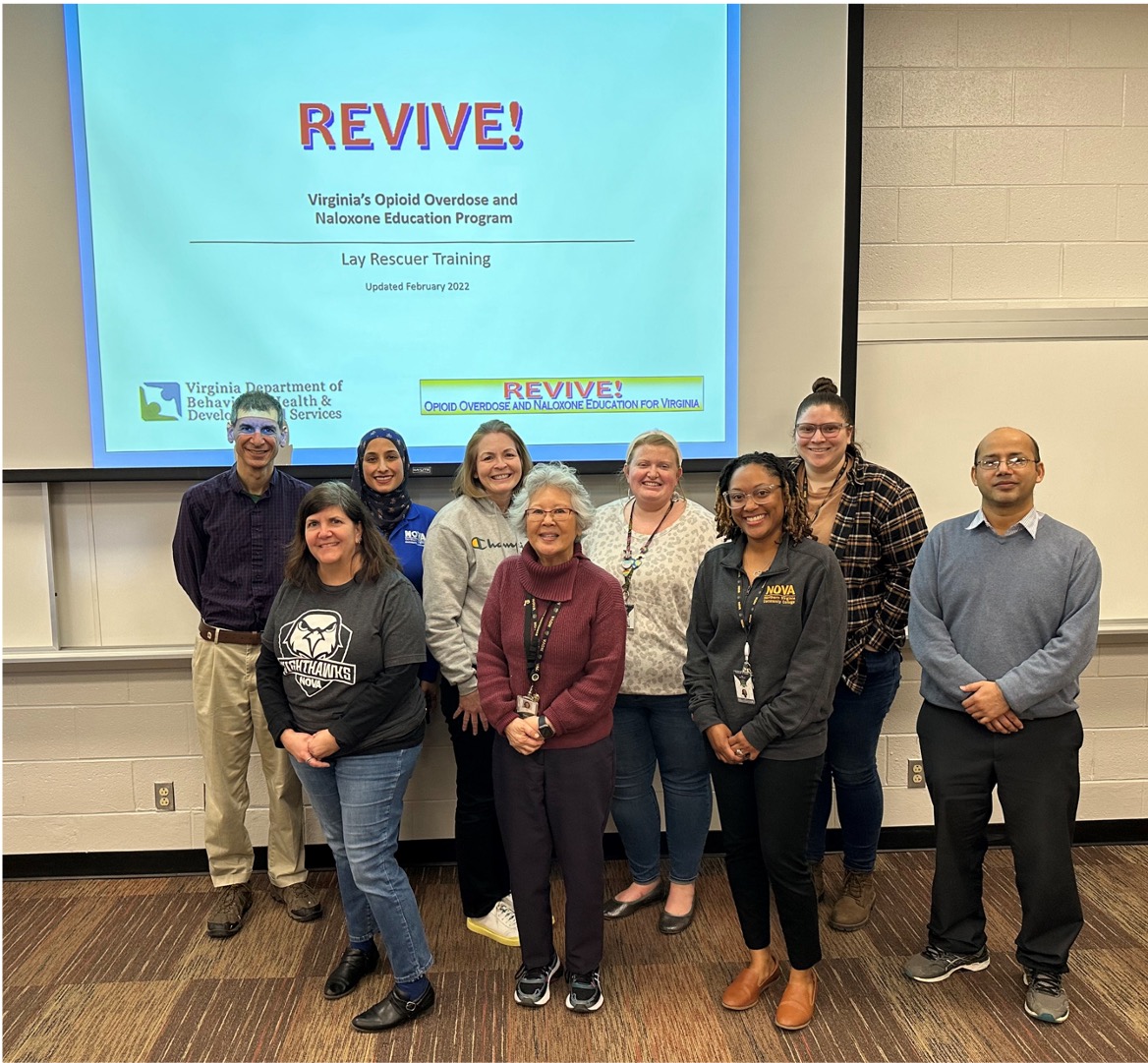 Participants in the REVIVE! training class at NOVA's Loudoun Campus pose for a photo in front of a screen at the front of a classroom. The projected slide reads as follows: "REVIVE! Virginia's Opioid Overdose and Naloxone Education Program. Lay Rescuer Training, Updated February 2022."
Pictured, from left to right, are Jeff Vetrano, Tiffany Blackmon, Saleha Bholat, Lisa Stelle, Lisa Fukui, Maddie Quick, Aja Adu-Gyamfi, Tori Rodriguez and Chola Chhetri.
