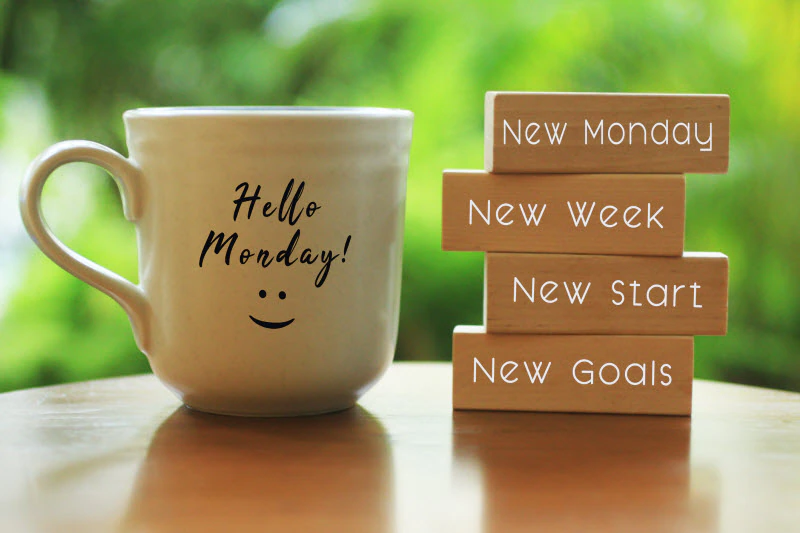 Photo. A cream-colored coffee mug with the words "Hello Monday!" and a smiley face sit on a table next to a stack of four Jinga blocks that say, "New Monday," "New Week," "New Start" and "New Goals." Spring green leaves are blurred in the background.