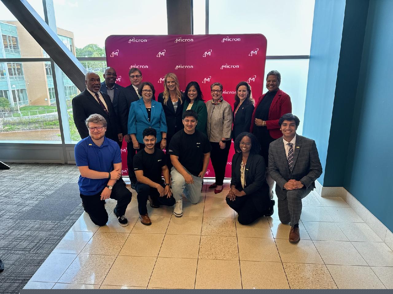 Front row: Five current and former NOVA students, two of whom are now Micron employees, joined officials, including Loudoun Campus Provost Dr. Julie Leidig (back row, fourth from left), Acting U.S. Secretary of Labor Julie Su (back row, fourth from right) and NOVA President Dr. Anne Kress (back row, third from right) at Micron’s announcement of a registered apprenticeship program in partnership with the College and the National Institute for Innovation and Technology. The group smiles for the camera in front of a bright pink step-and-repeat with windows looking out on NOVA's Loudoun Campus behind.