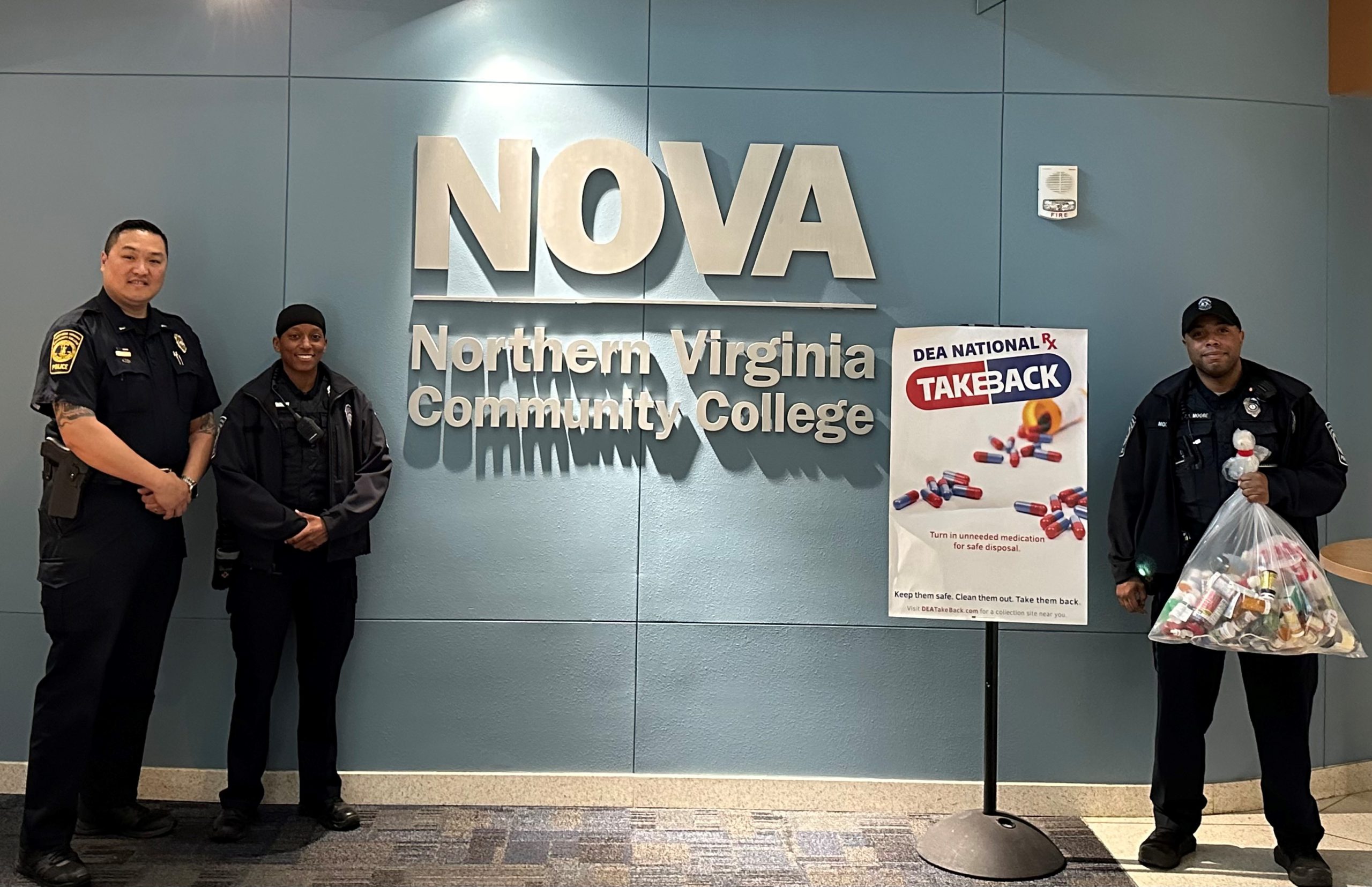 Photo of NOVA Police officers in front of silver NOVA sign in the Student Services building on the Annandale Campus. Lieutenant Tony Ong and Officer Cherelle Rowe appear at left, while Officer Jarrin Moore, next to a sign advertising the DEA Take Back Day event and holding a clear bag of unused and expired prescription medications, appears at right. 