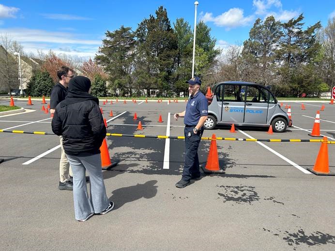 Photograph of two NOVA community members listening to a police officer discuss the cone course set up in a parking lot on NOVA's Loudoun Campus for a distracted driving awareness event. A silver vehicle resembling a golf cart and emblazoned with "Virginia State Police" in blue sits in the background. 