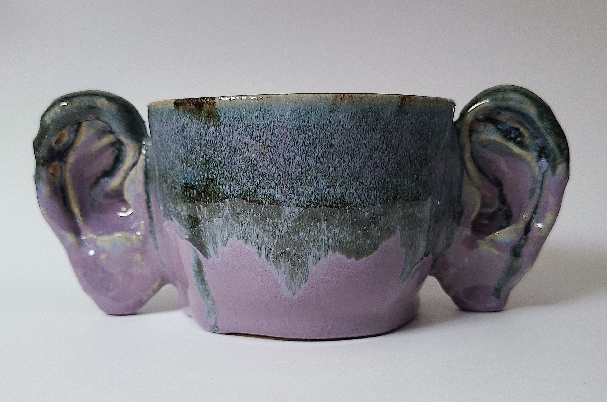 Photograph of small purple ceramic vessel titled “Ear Mug #4” by Annandale student Lee Soderborg. The vessel, which resembles a drinking mug or cup, is glazed in shiny purple and gray tones and has two handles in the shape of human ears. 