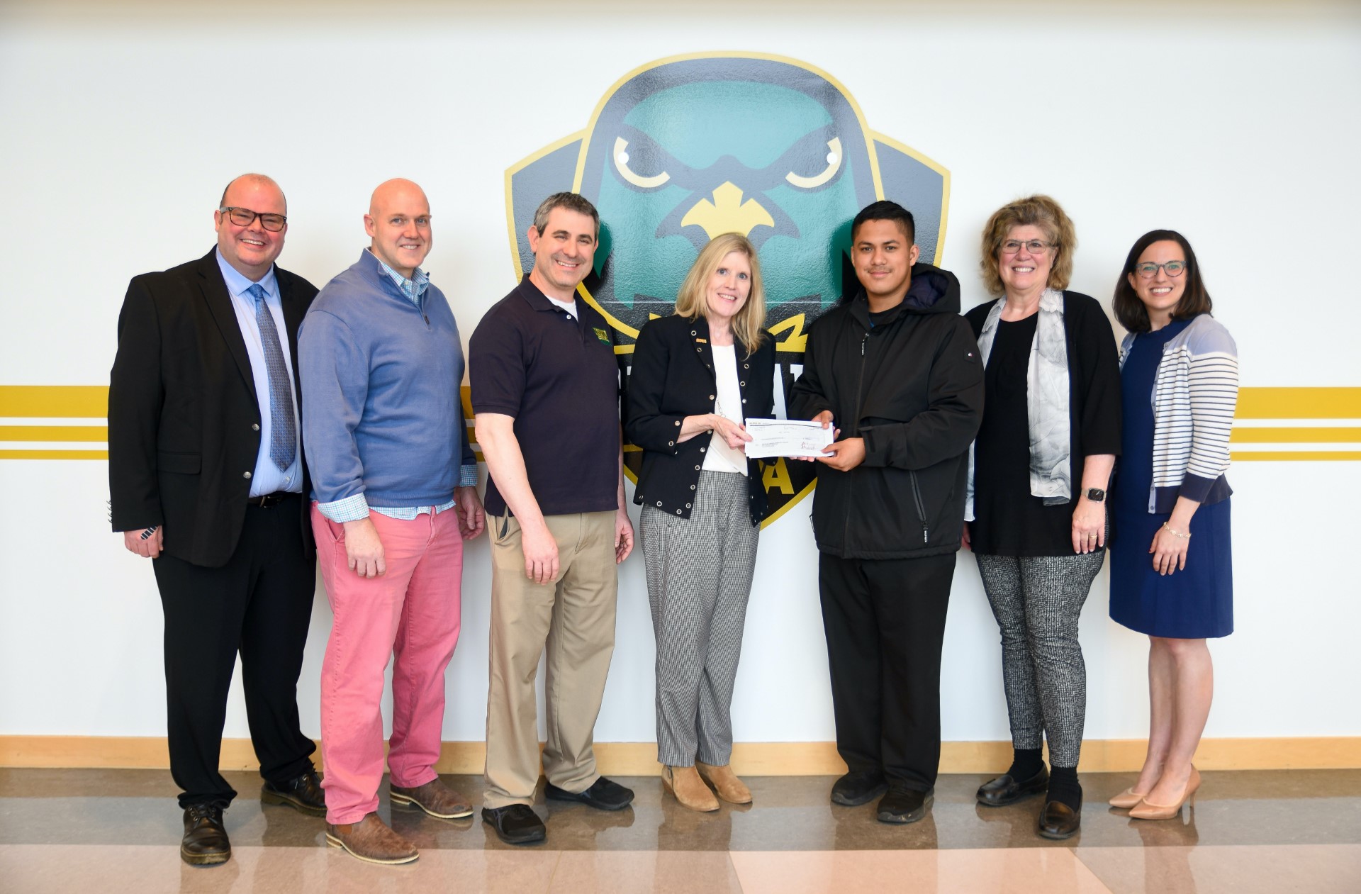 Event attendees at the check-presenting ceremony pose for a picture in front of a large decal of Ace the Nighthawk. From left to right: Bryan Brown, Chair, NOVA Cinema Program; Chris Stallings, Professor, NOVA Cinema Program; David Epstein, Academic Dean, Woodbridge Campus; Kelly Persons, Executive Director, NOVA Foundation, Angel Pino, student and Phi Theta Kappa member; Theana Kastens, Honors English Faculty and advisor to PTK's Woodbridge chapter; and Abbey Jones, Associate Director, Specialty Programs.