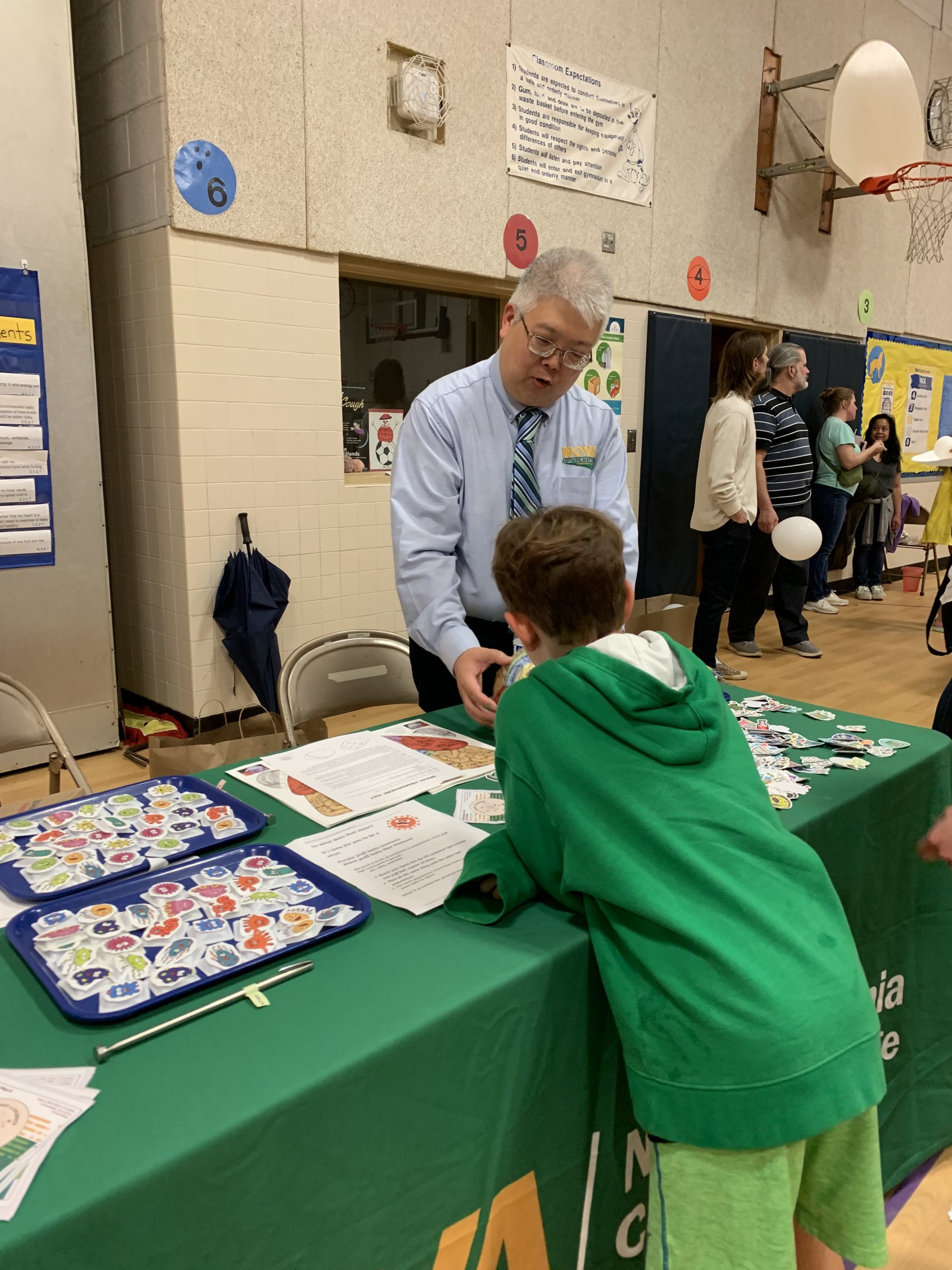 Table draped with a green NOVA cloth and containing free stickers for children to take. Annandale Chemistry Professor Ben Wang impresses a child with science fun facts. 