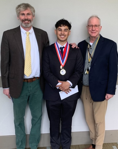 Javier Sagastume beams while wearing his medal on a red, white and blue ribbon. He is flanked by PTK Co-Advisors. On left, Stephen Clarke and, on right, Christopher Blake. 