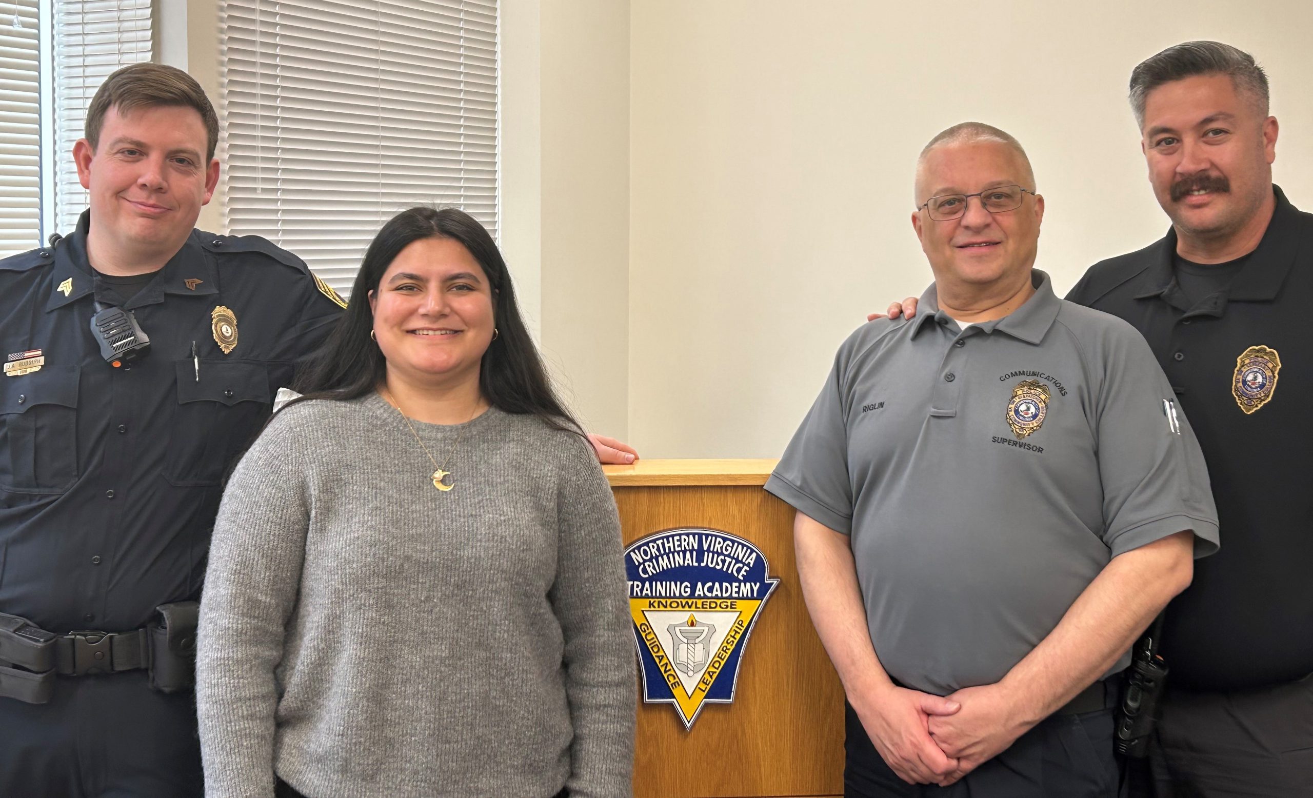 Photograph of NOVA PD with new dispatcher. At left of podium draped with "Northern Virginia Criminal Justice Training Academy" flag are Sergeant Jeffrey Rudolph and Dispatcher Sahar Baluch. At right of podium are Dispatch Supervisor David Riglin and Sergeant John Baxter.