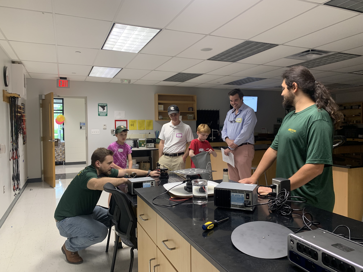 Professor Alex Bohn (kneeling) one of our physics professors demonstrates how to visualize sound in an artistic way using a Chaldni plate as Prof. Nathaniel Green (wearing a DC United Ballcap) and his son, another dad and his son and a NOVA student volunteer look on.