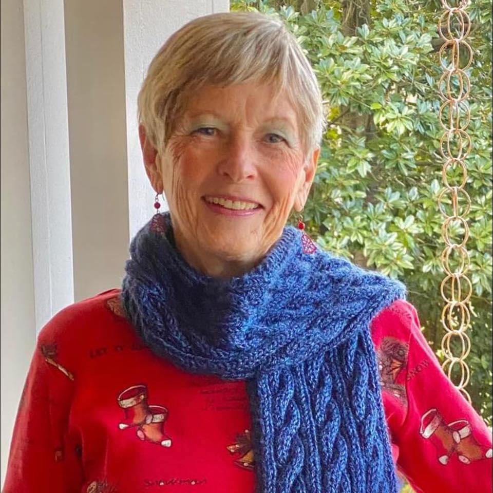 Jane Martin, NOVA alum and member of the College's inaugural nursing class, smiles for the camera in front of a window. She has short light hair and wears a red printed blouse with a cable-knit blue scarf around her neck.