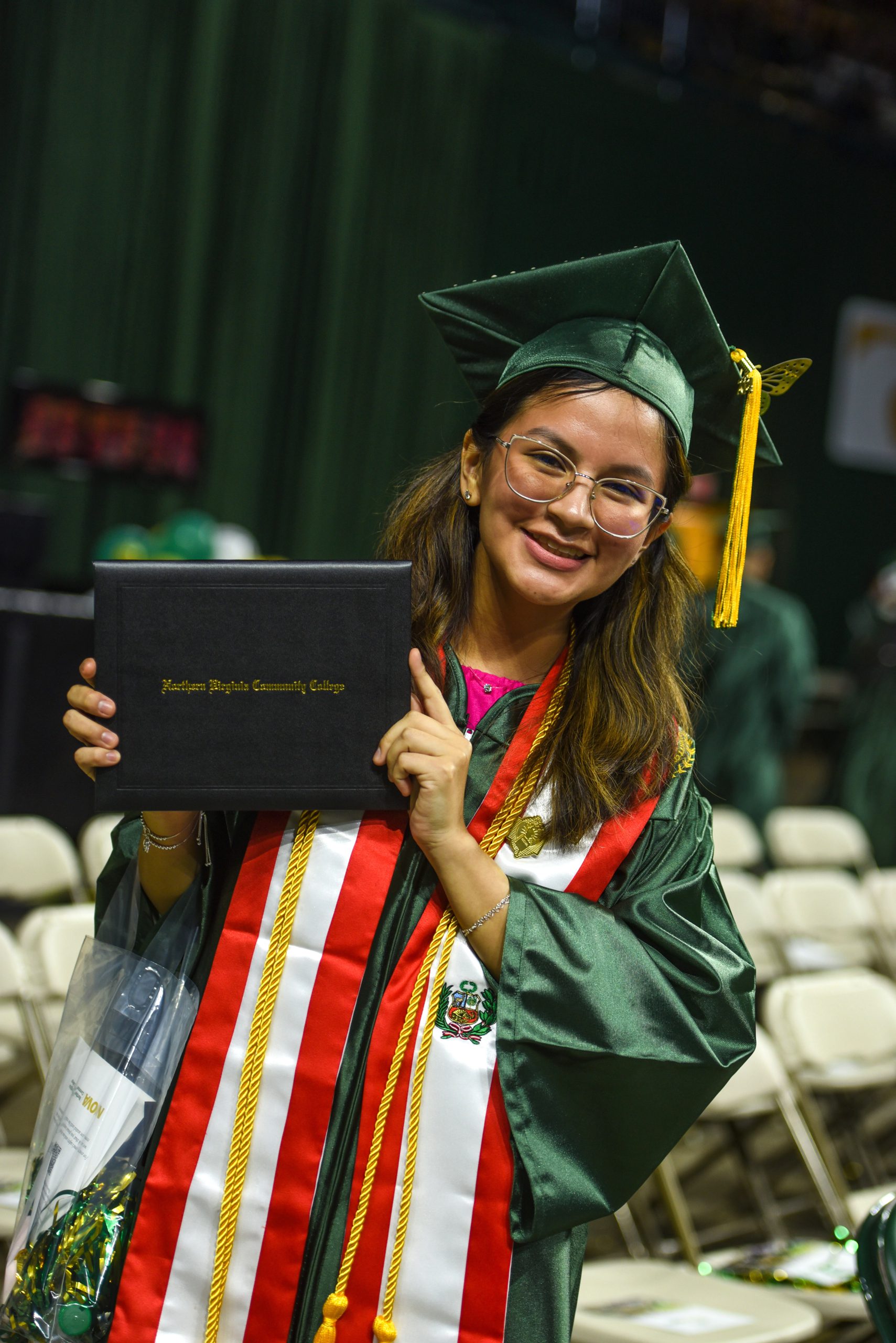 JKC Foundation Undergraduate Transfer Scholarship winner and NOVA Nighthawk Catherine Ercilla smiles for the camera while holding her diploma! She wears a green robe, green mortarboard with gold tassel, and different honor cords.