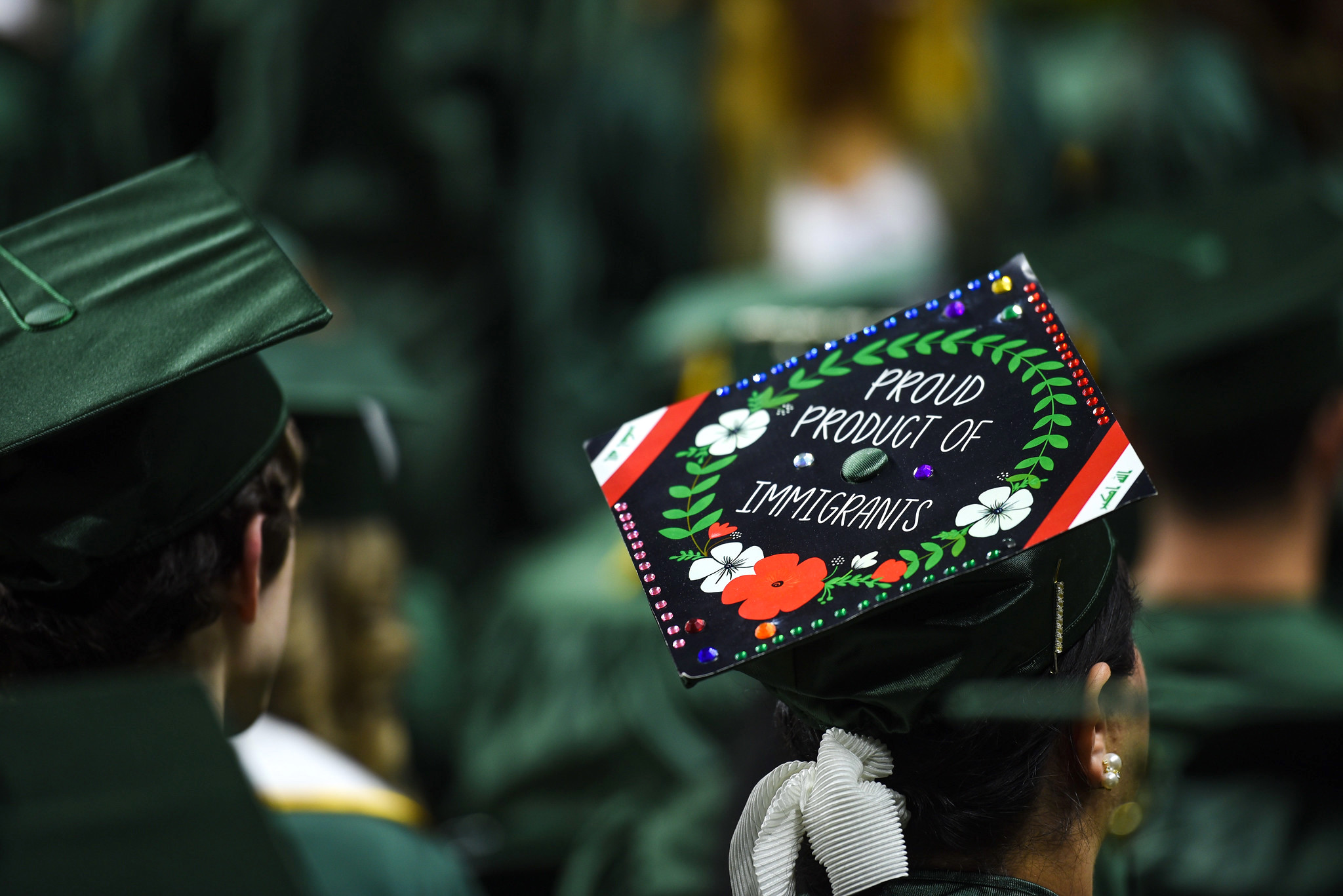 Nour's brightly decorated motar board with a bright wreath in green with white and red flowers and the words "Proud Product of Immigrants." 