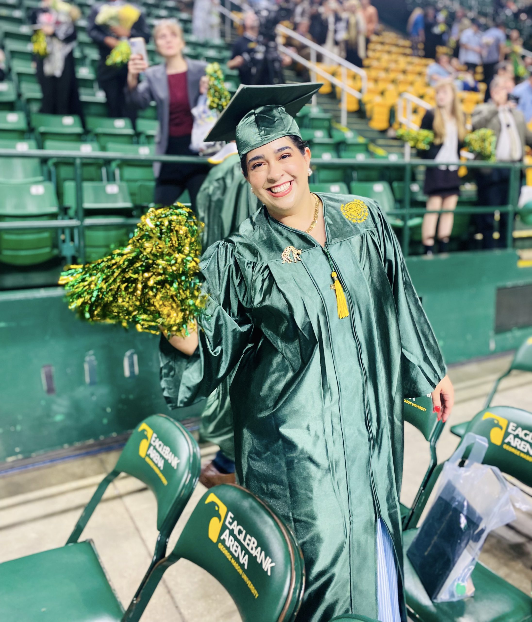 Nour Alamiri, dressed in her graduation regalia waves a green and gold foil pom pom in celebration of her big day!