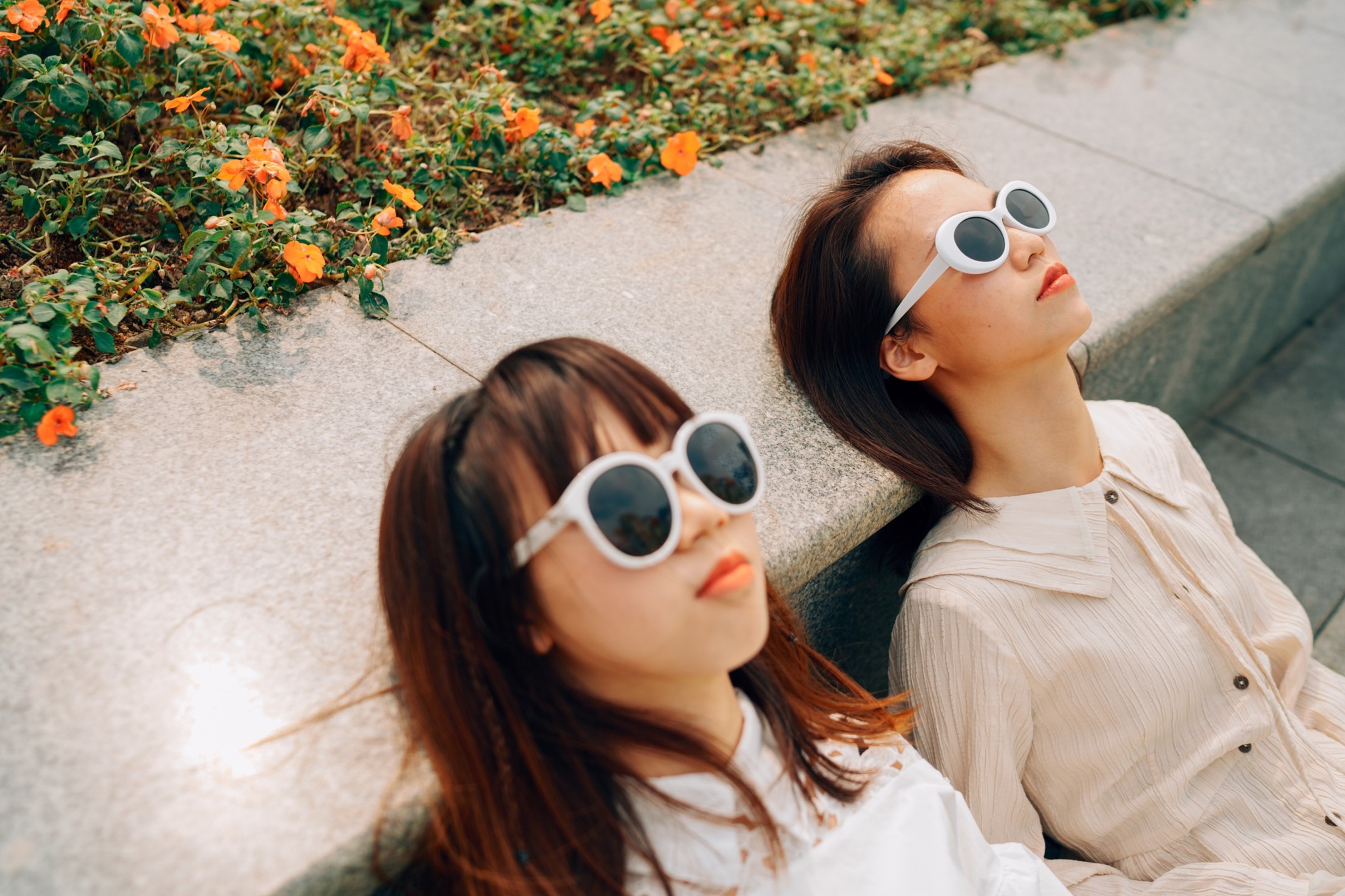 Two young women wearing white shirts and white sunglasses take a break, leaning their heads against a concrete stoop with orange flowered vinca vines in a flower bed behind them