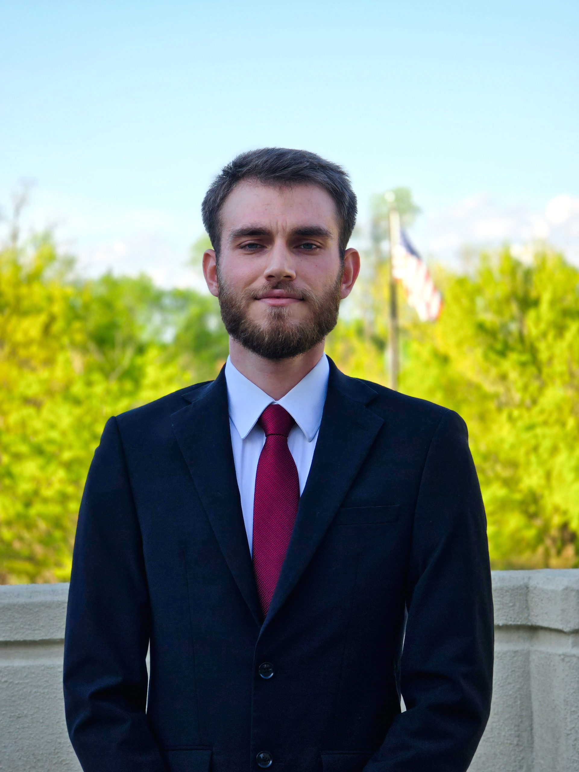 Tariq Aldalou, soon-to-be NOVA graduate, smiles while outside on a sunny day. He wears a dark suit, white shirt, and red tie, and an American flag flies in the background among the green treetops.