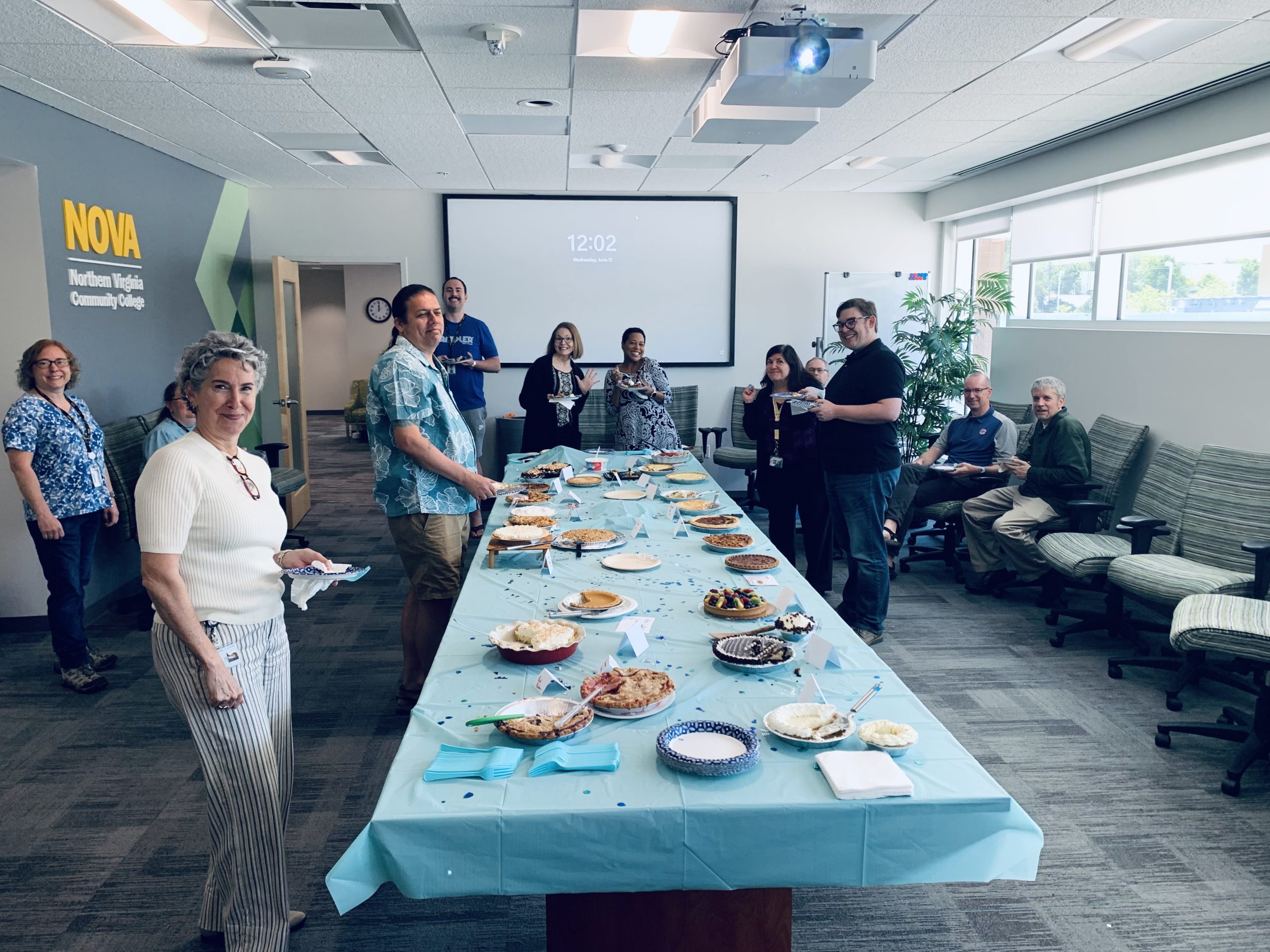 Photograph of NOVA Loudoun faculty and staff gathered in a conference room to celebrate Pie Day on June 12, 2024. In the center of the photo is a long table, covered in a light blue tablecloth, laden with various sweet and savory pies. A number of people stand at the side of the table and behind it to smile for the camera. 