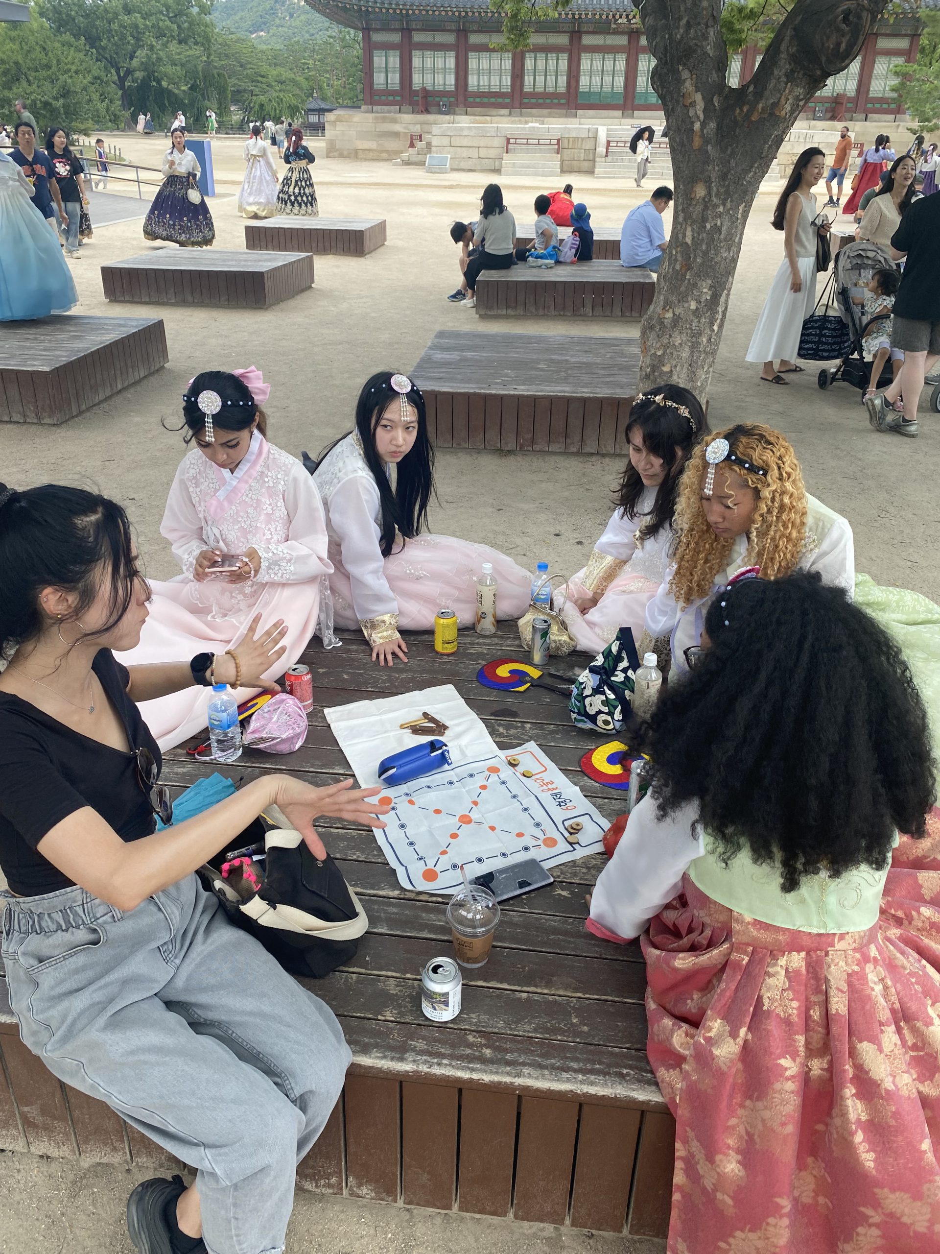 A group of six young women, several in korean traditional dresses, play Yut Nori in a shaded courtyard.
