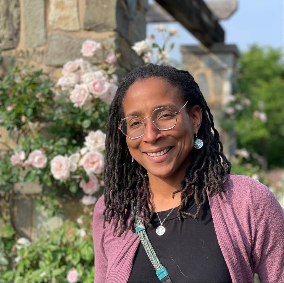 Melanie Jenkins smiles while standing in front of a beautiful rock wall with a trellis of pink roses.