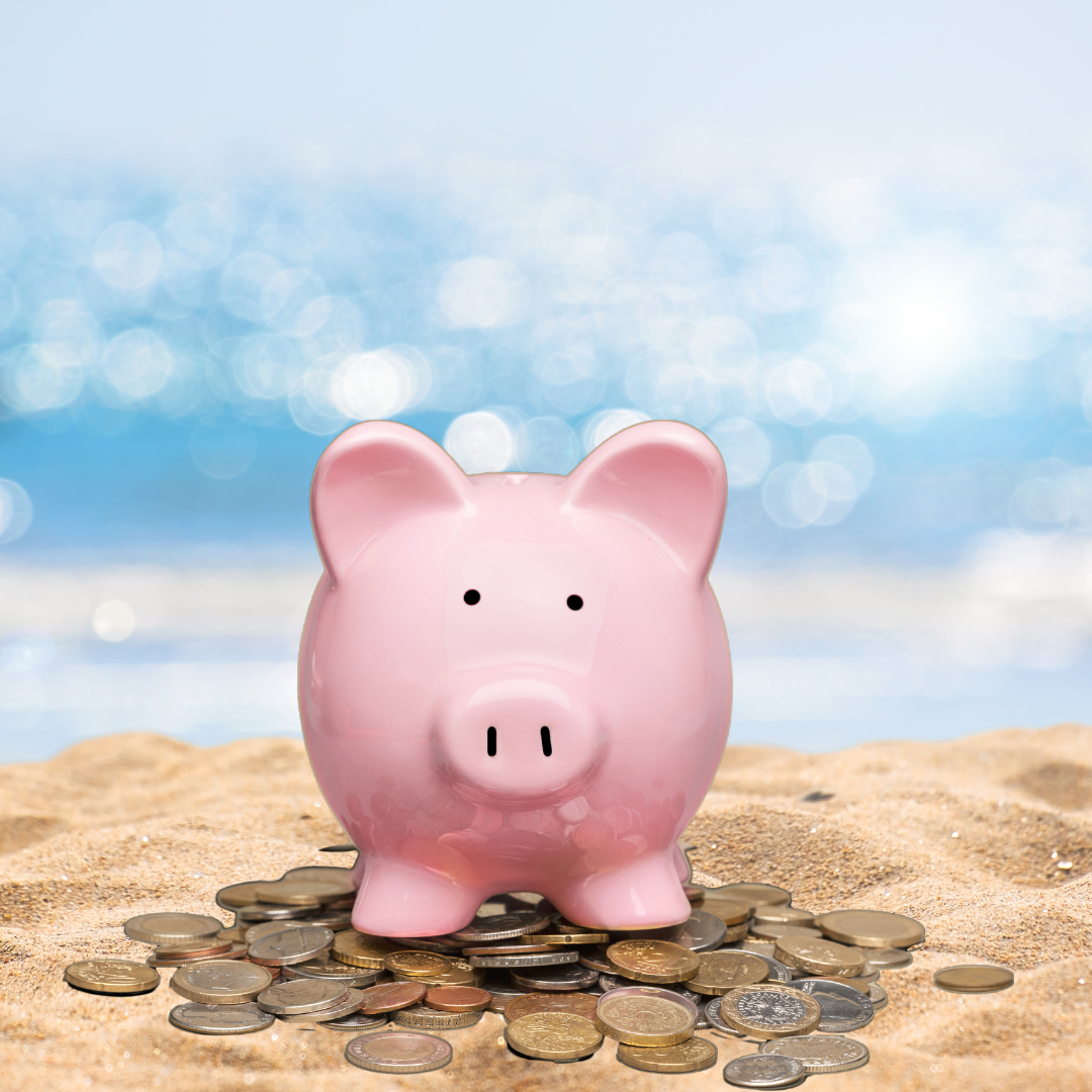 Piggy bank stands on the sand on top of a handful of coins. The ocean glimmers in the background