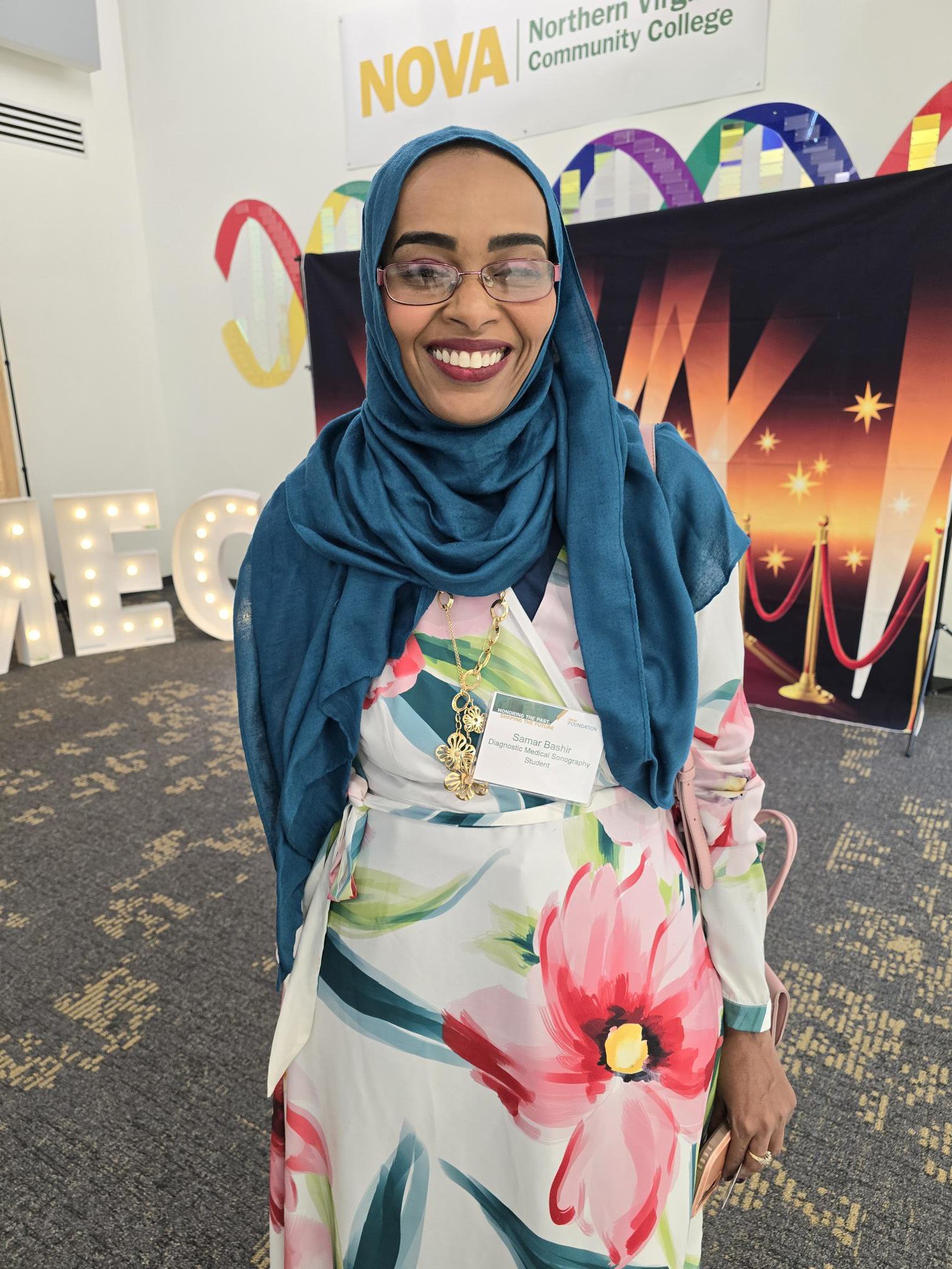 Sonography Student Samar Bashir smiles brightly as she stands in the MEC first-floor Forum wearing a beautiful flowered dress and blue headscarf.