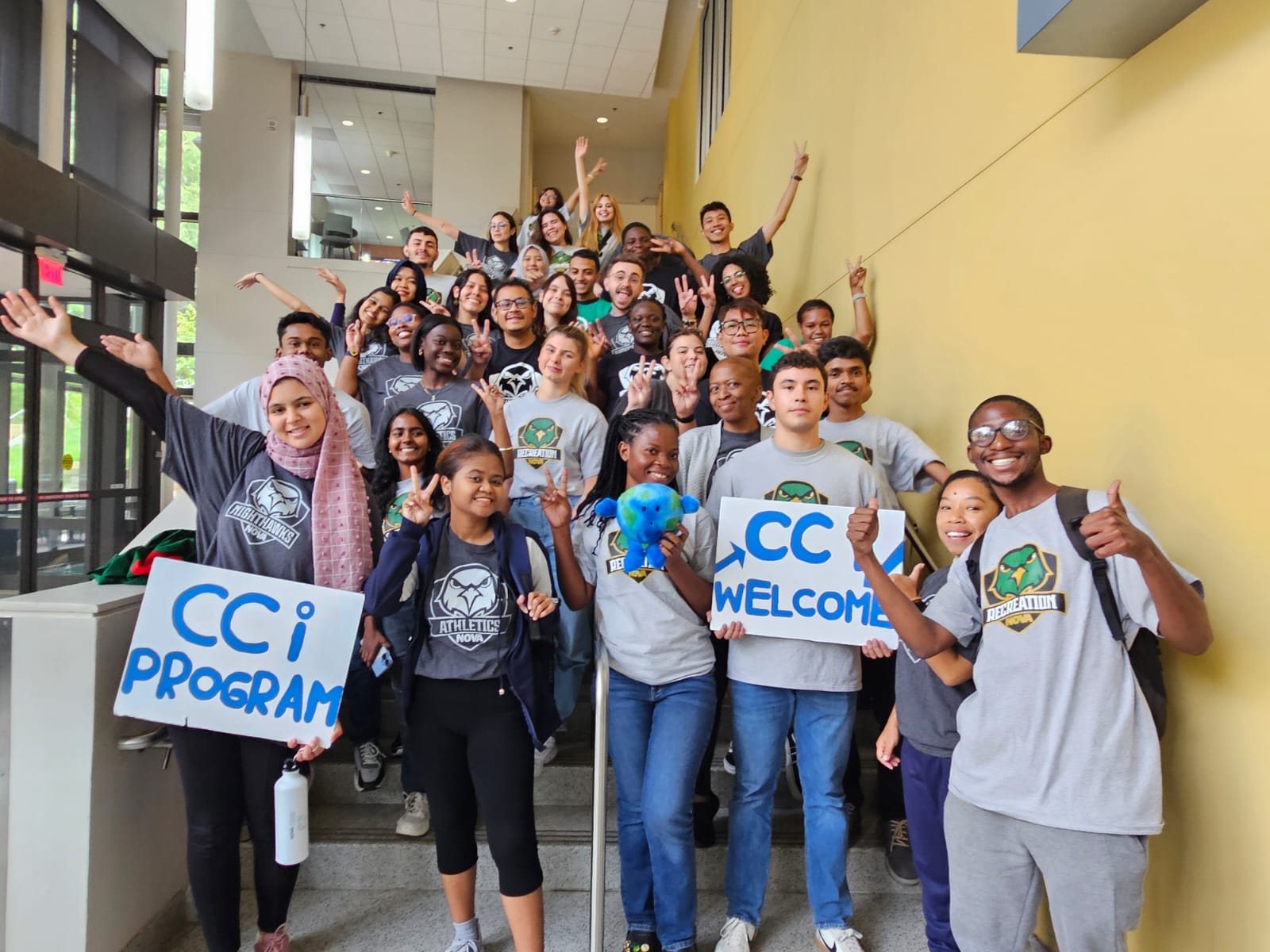 CCI students line up on the stairs outside the Annandale Campus Bookstore, waving, giving the peace sign and holding signs that say CCi Program and CCI Welcome! One holds a stuffed animated globe. 
