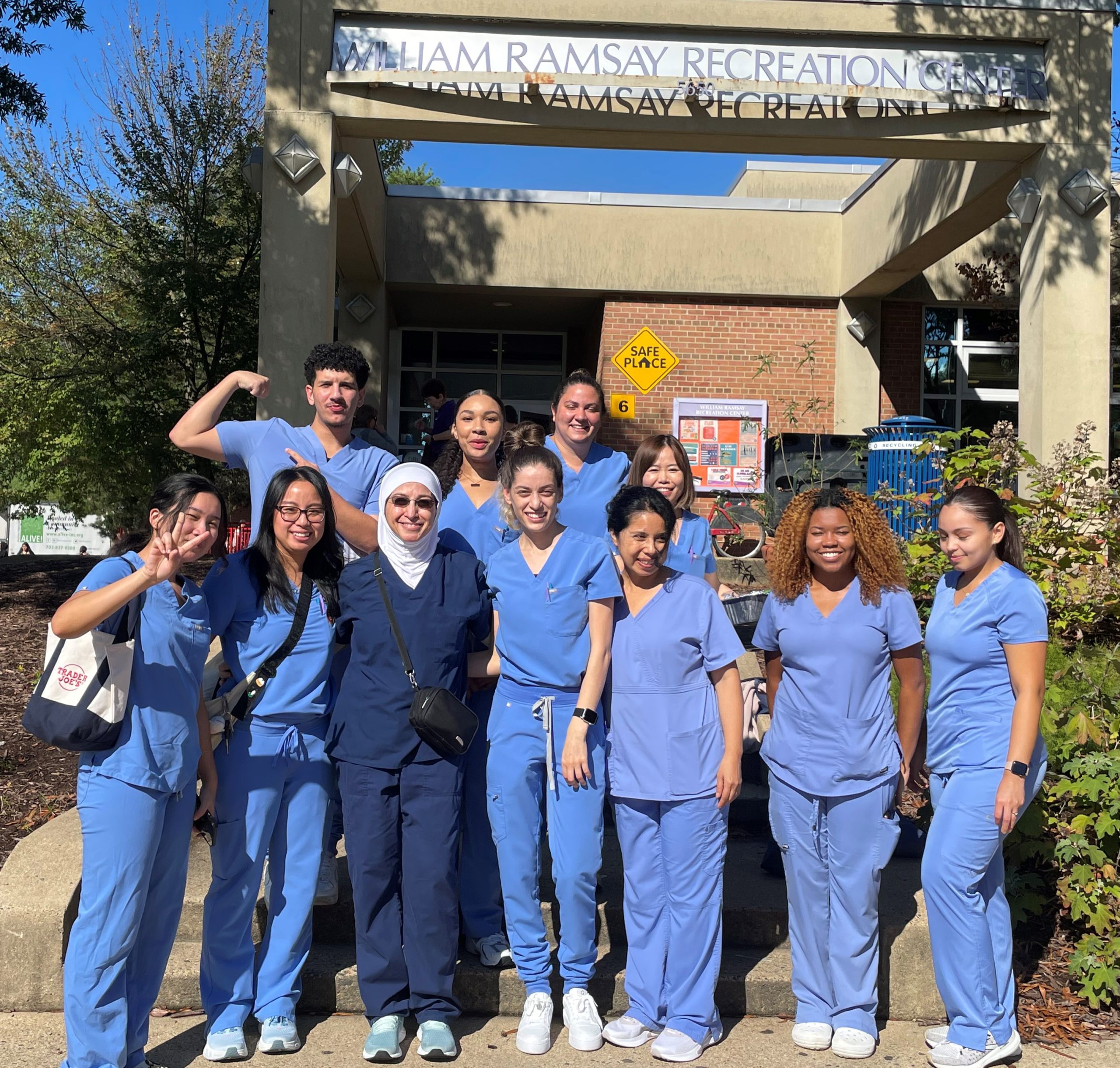 Ten NOVA Dental Hygiene students, dressed in medium-blue scrubs stand with their instructor in navy blue scrubs and a white hijab in front of the William Ramsay Recreation Center.
