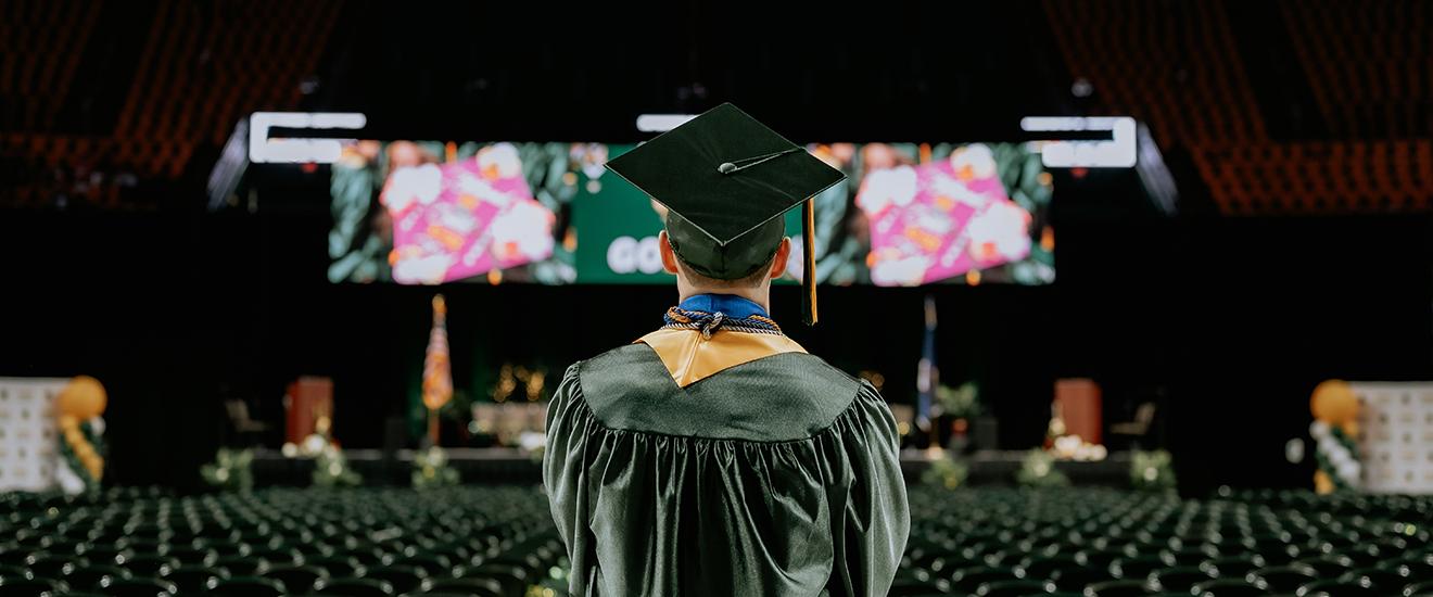 Graduate wearing green regalia, gold sash and mortarboard looks toward the graduation stage
