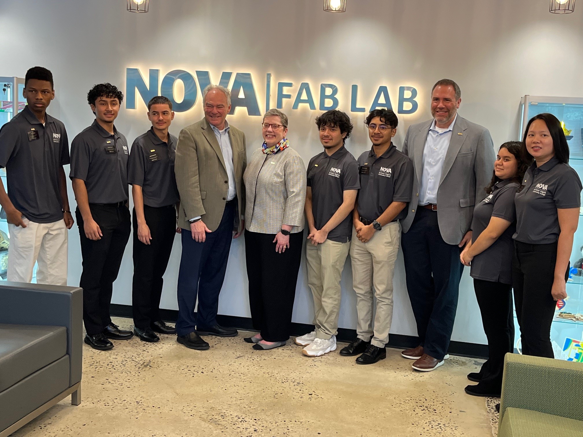 Senator Tim Kaine (4th from Left) poses with a group of students and Dr. Anne Kress (5th from Left) and Dr. Chad Knights 3rd from Right) beneath the backlit letters that read NOVA Fab Lab.