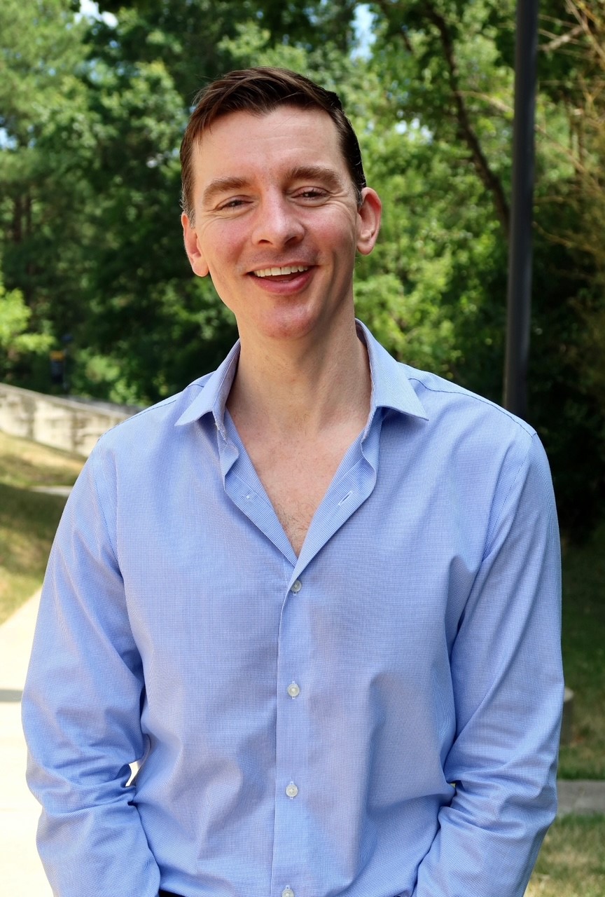 Portrait of Ryan Thomas Finnegan standing outdoors on a beautiful spring day with trees and a white splitrail fence in the background.