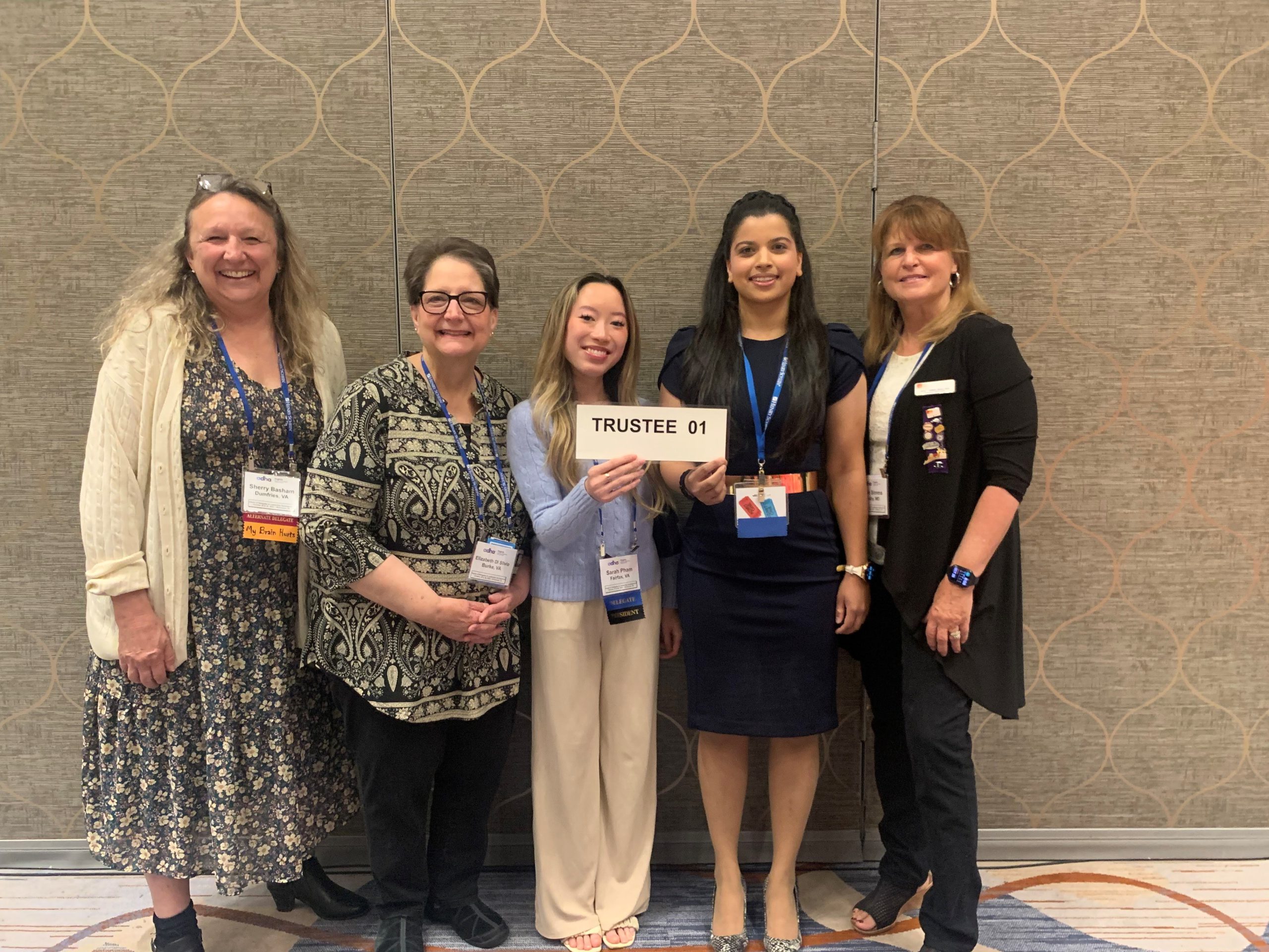 Full-Time Dental Hygiene Faculty Sherry Basham and Betsy Di Silvio, Students Sarah Pham and Shalini Jain and Adjunct Julie Simms smile for a photo. Sarah and Shalini hold up a sign that reads "Trustee 01"