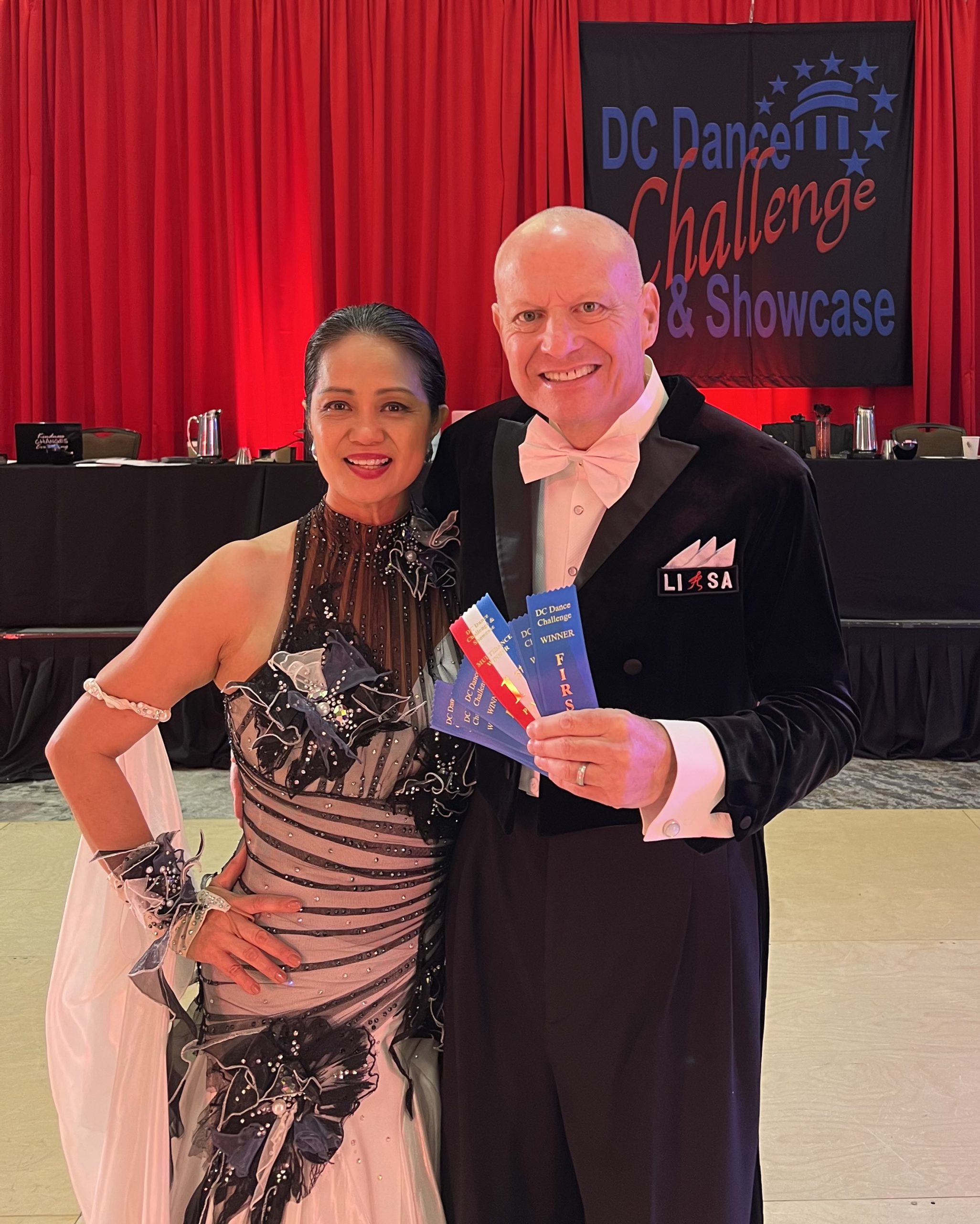 NOVA Dance Instructor Alan Golombek and his wife, Thuy, at the DC Dance Challenge this past weekend. Alan, at right, wears a black tuxedo with white shirt and tie. Thuy, at left, wears a silver, black, and white gown, with her held pulled back in an elegant updo. The couple proudly displays their blue ribbons while smiling for the camera!