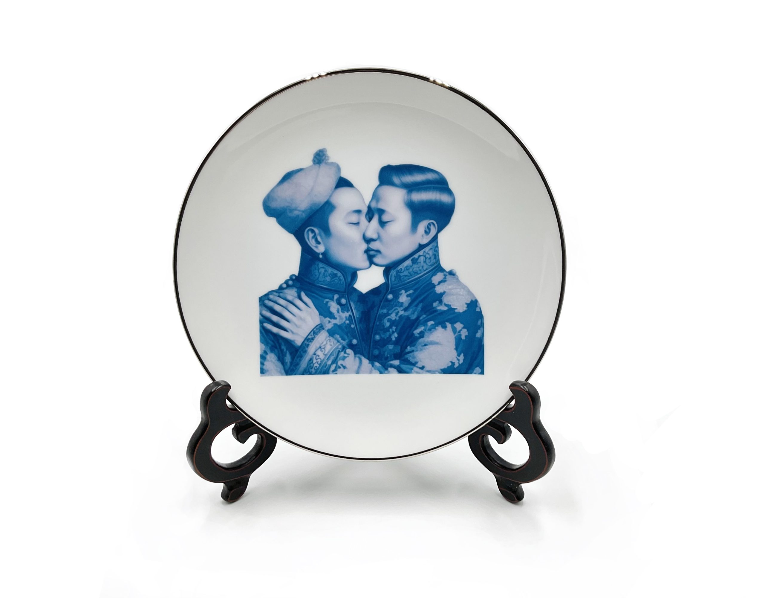A white porcelain plate with a black painted edge and a blue image in the center with two Asian people kissing. The one man has his hand on the other's shoulders and the hand has a thumb and five fingers.