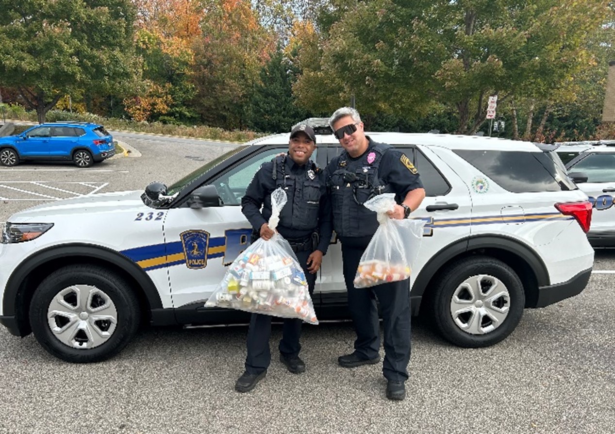 Officer Jarrin Moore (left) and Sergeant John Baxter stand next to a NOVA police cruiser holding bags of medications collected for proper disposal.