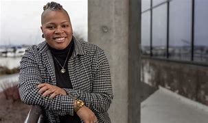 Charity Blackwell smiles for a picture as she leans against a railing wearing a black and white houndstooth blazer, a black turtleneck, and a silver cross necklace layered on a silver circular necklace with the city skyline behind her.