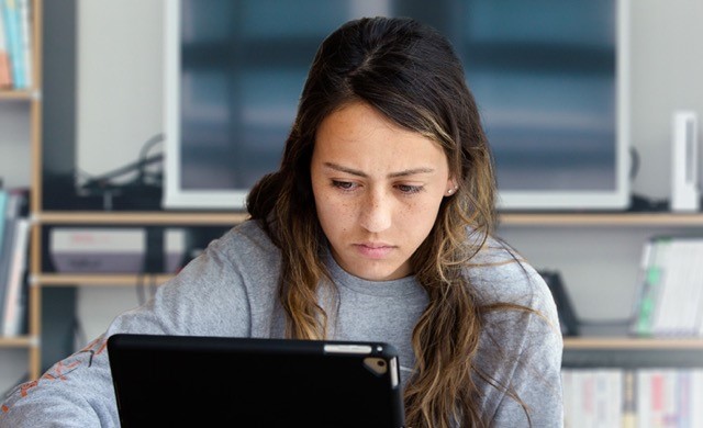 NOVA Online student works on her laptop with a TV on a shelf behind her.